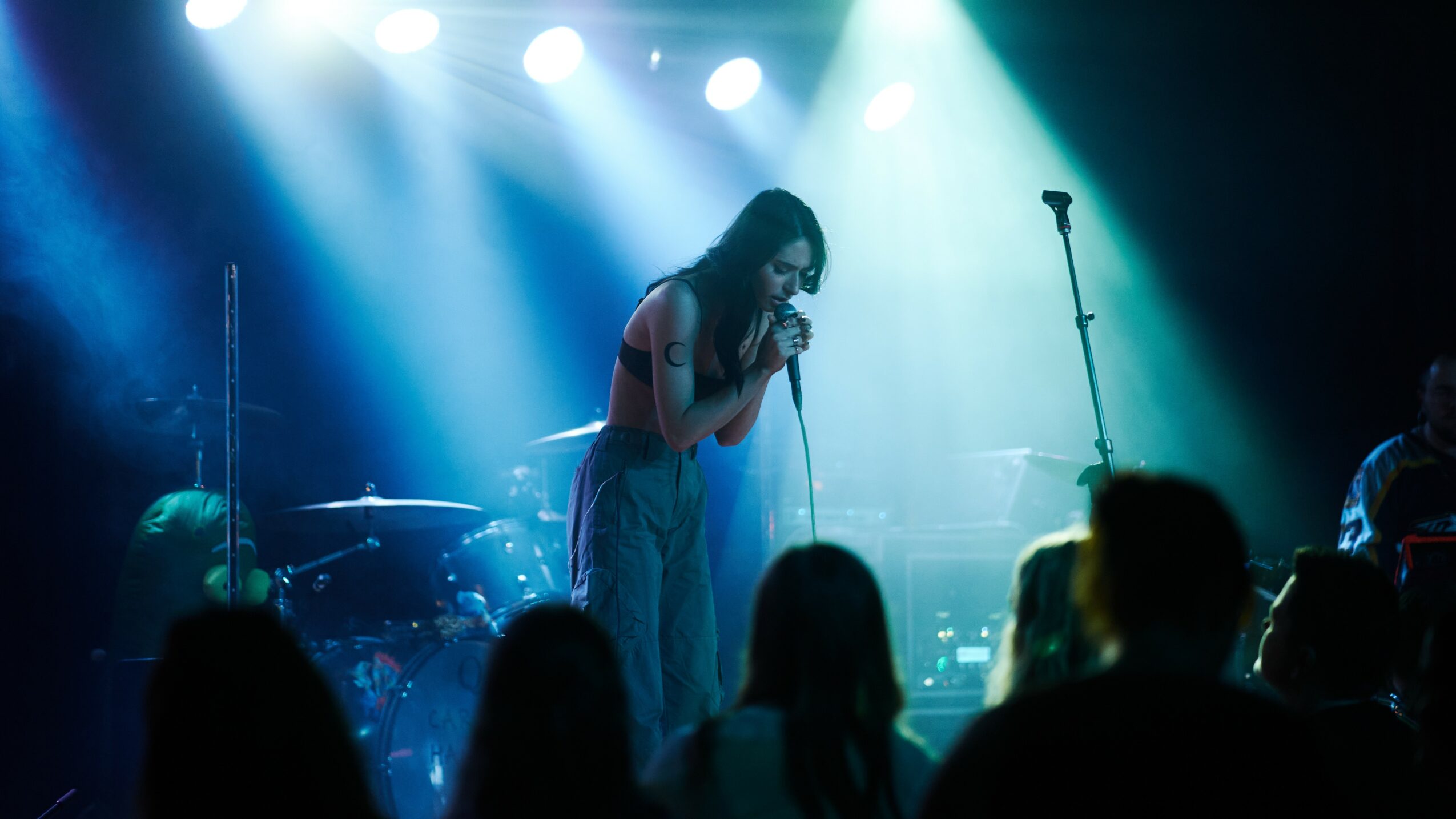 A woman sings into a microphone on stage with blue and green lighting, audience silhouettes visible in foreground.