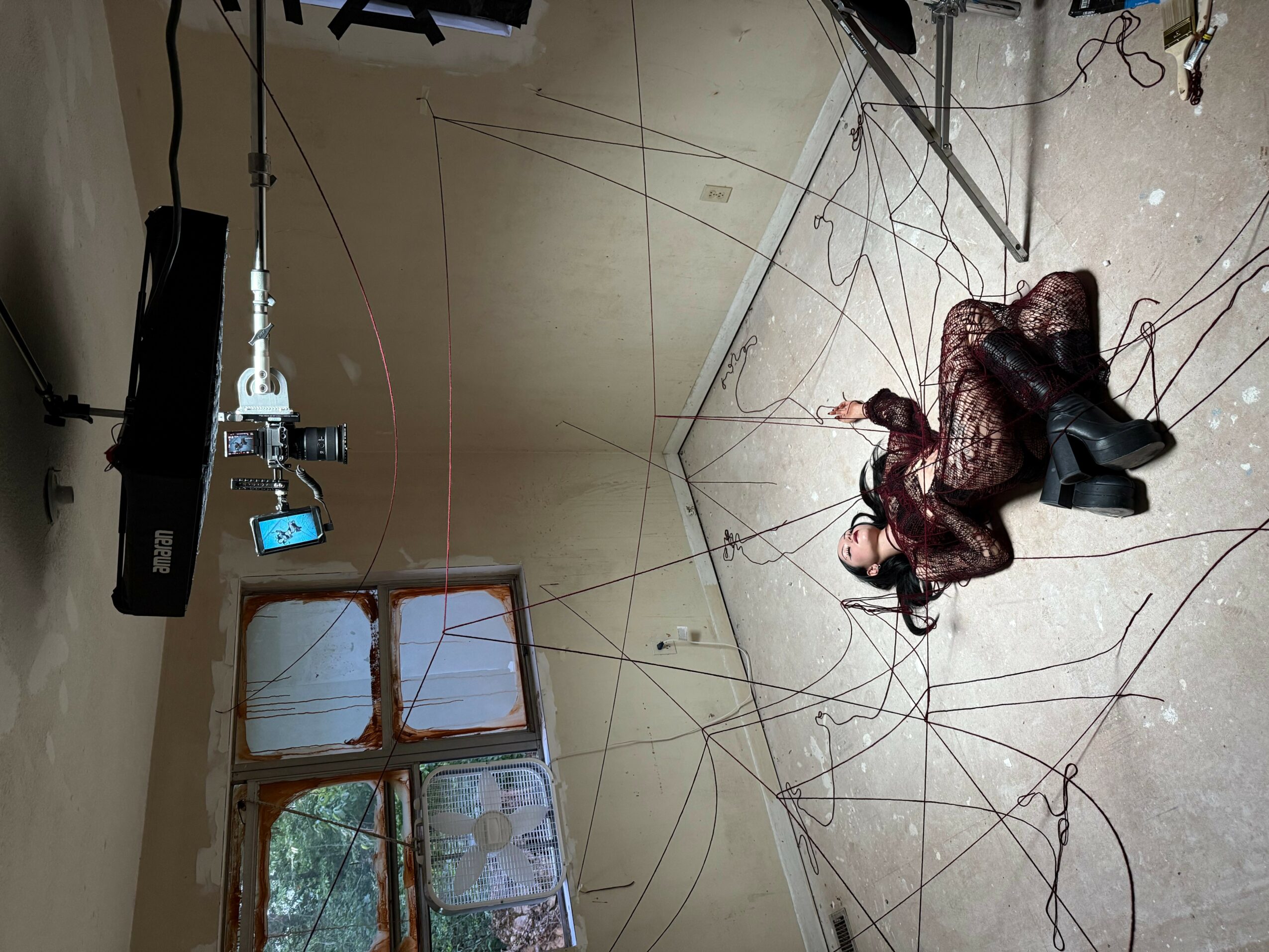 A person lying on the floor with long hair, surrounded by wires and equipment, in a room with a window.