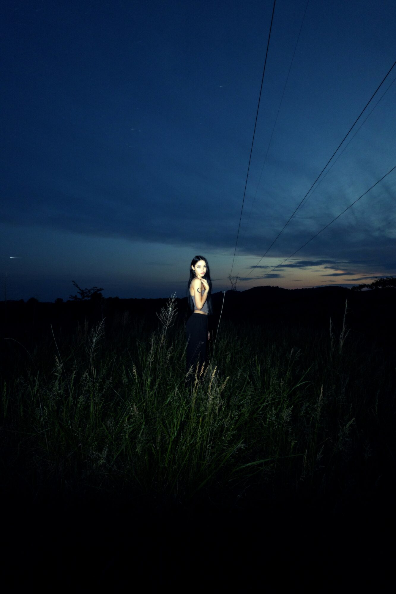 A person stands in tall grass under a dark, cloudy sky with power lines overhead at dusk.