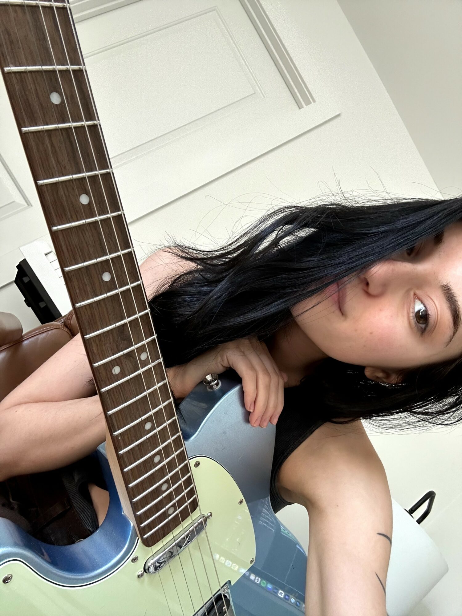 Young woman with long black hair holding an electric guitar, indoors with white walls and door in background.