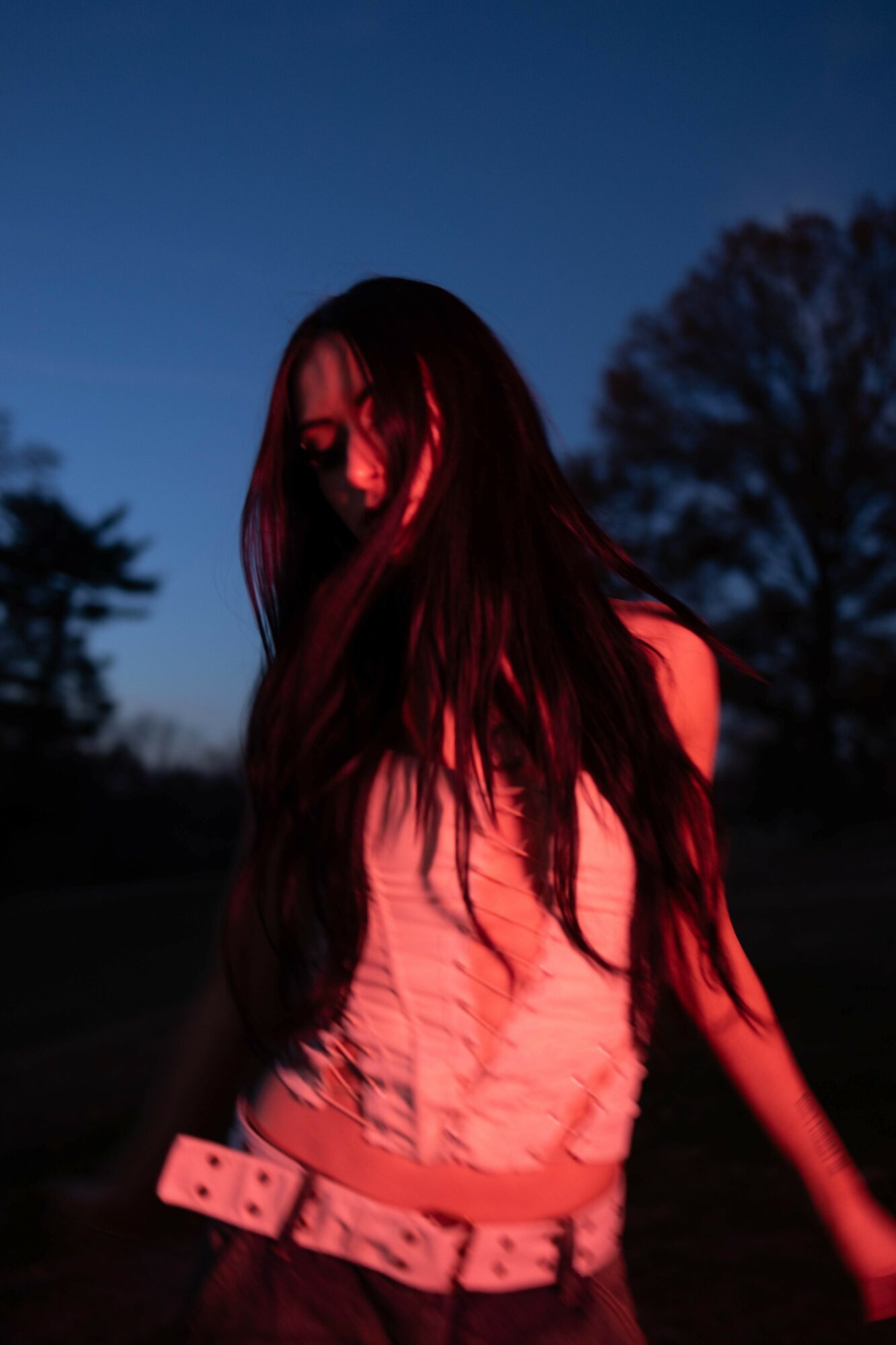 Person with long hair standing outdoors during dusk, illuminated by red light, with trees in the background.