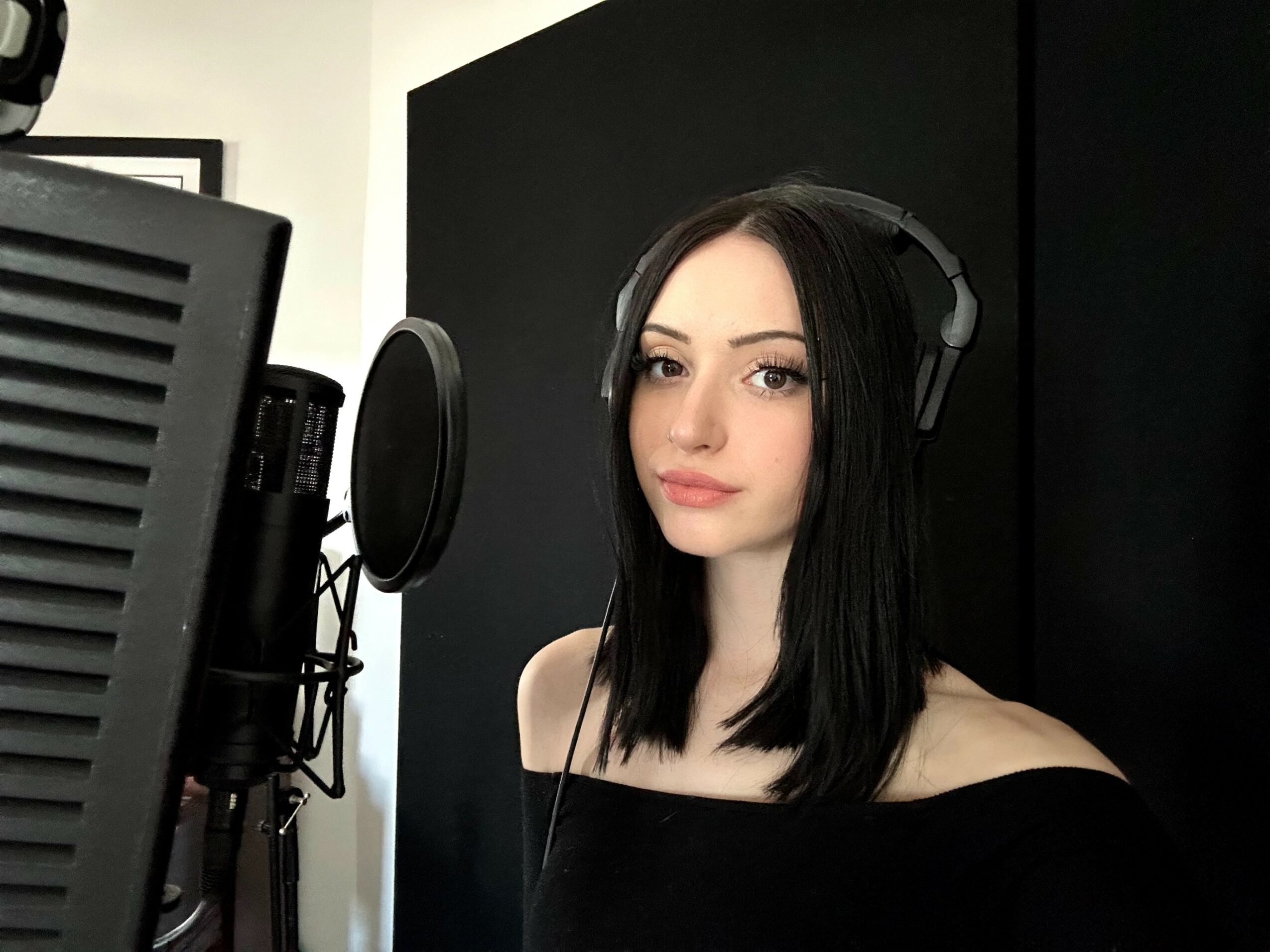 Young woman with black hair wearing headphones, standing in front of a microphone, black background, looking at camera.