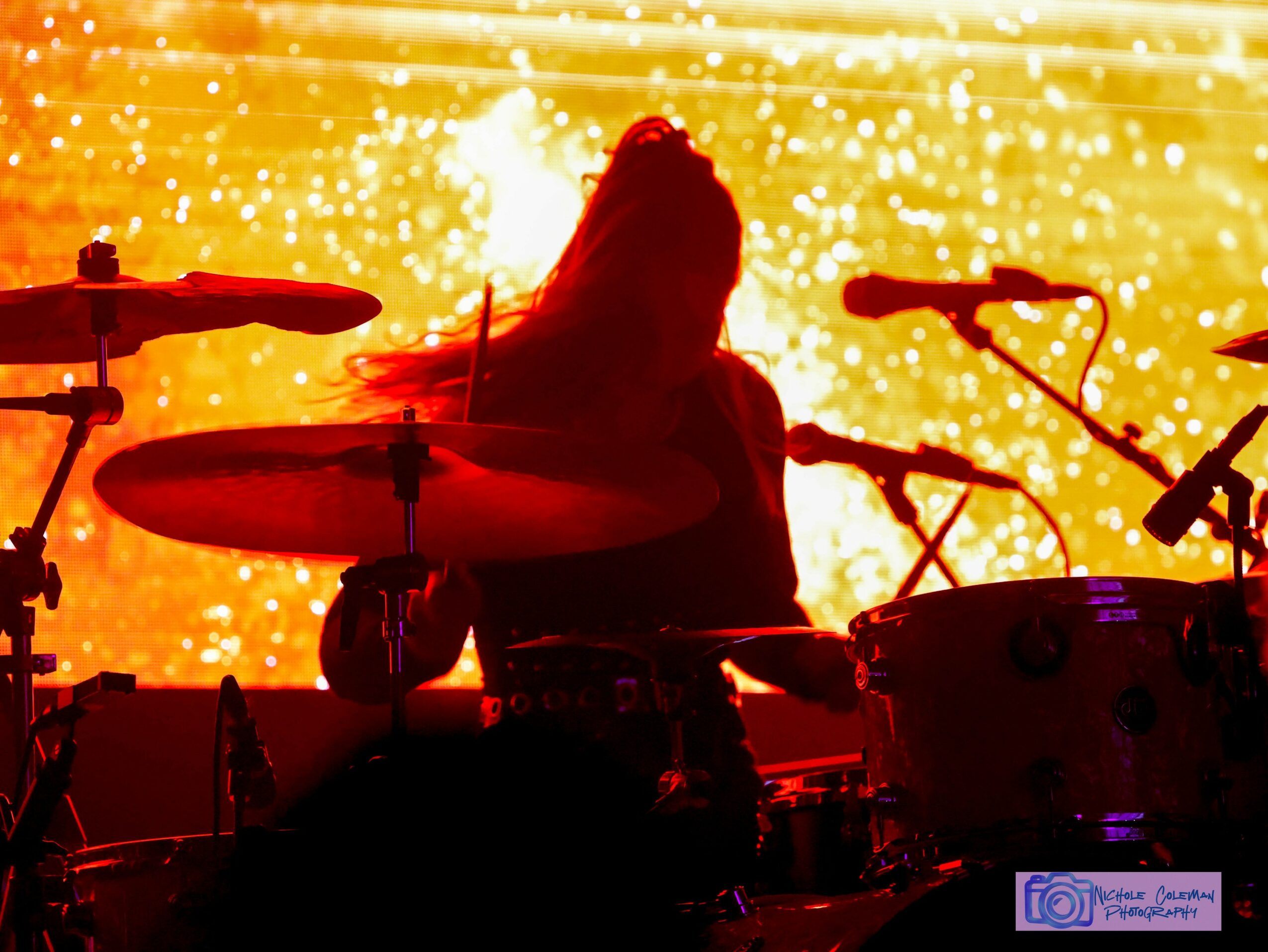 Silhouette of a drummer playing on stage with bright yellow and orange background, drums and microphones visible.