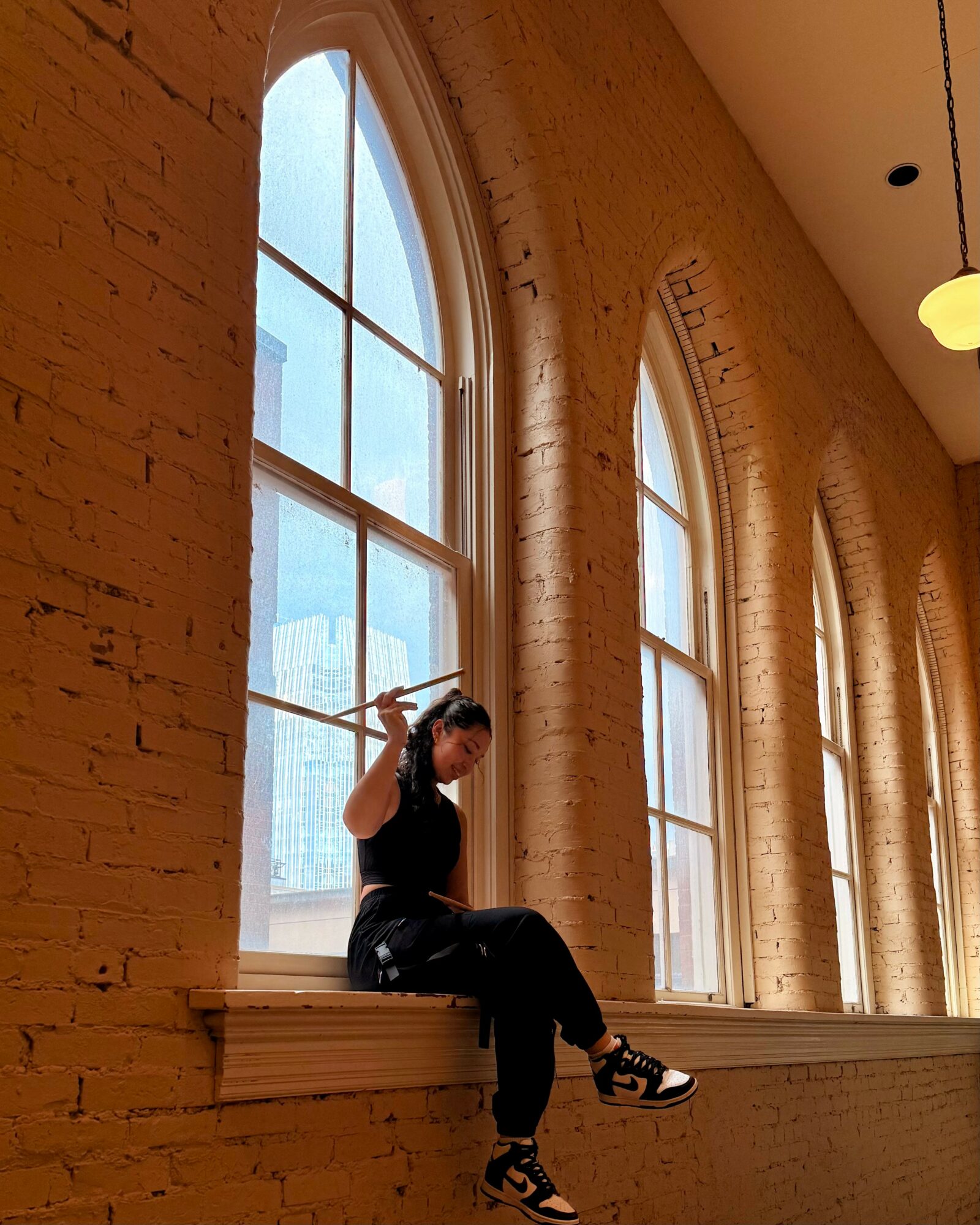 Person sitting on a windowsill inside a brick building, holding a camera or phone, with large arched windows behind them.