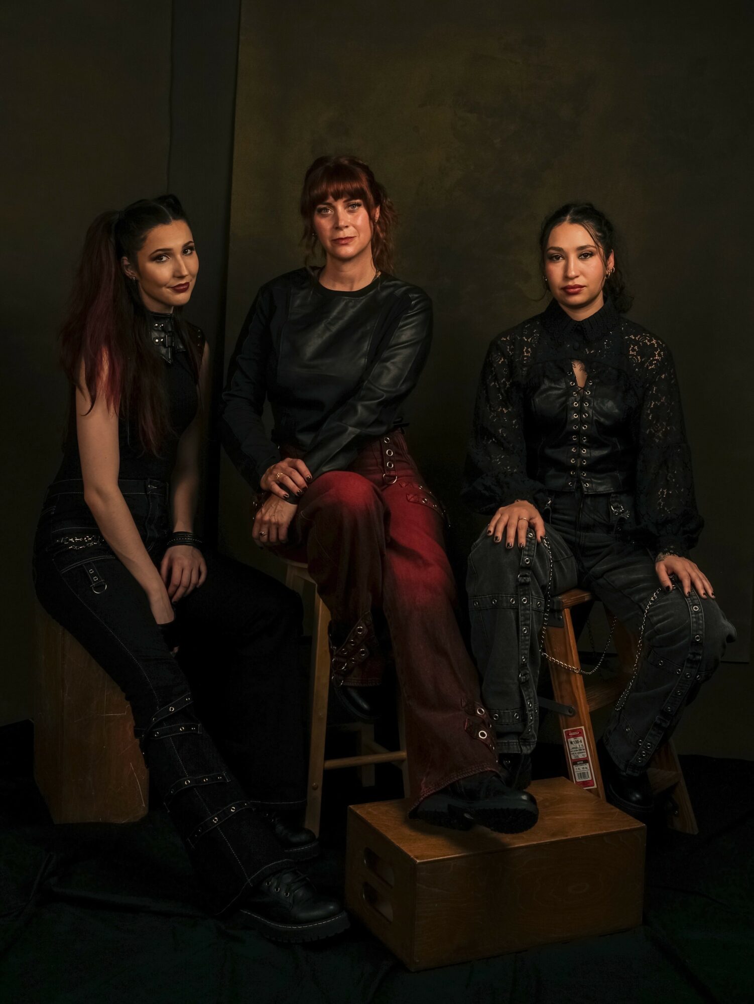 Three women sit on stools and a box against a dark background, looking at the camera. One woman has long dark hair, another has red hair, and the third has dark hair tied back.