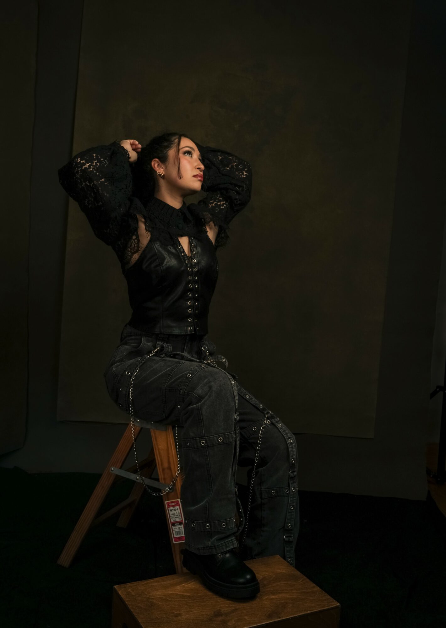 Woman sitting on a wooden stool with hands behind head, wearing dark clothing, in a dimly lit room.