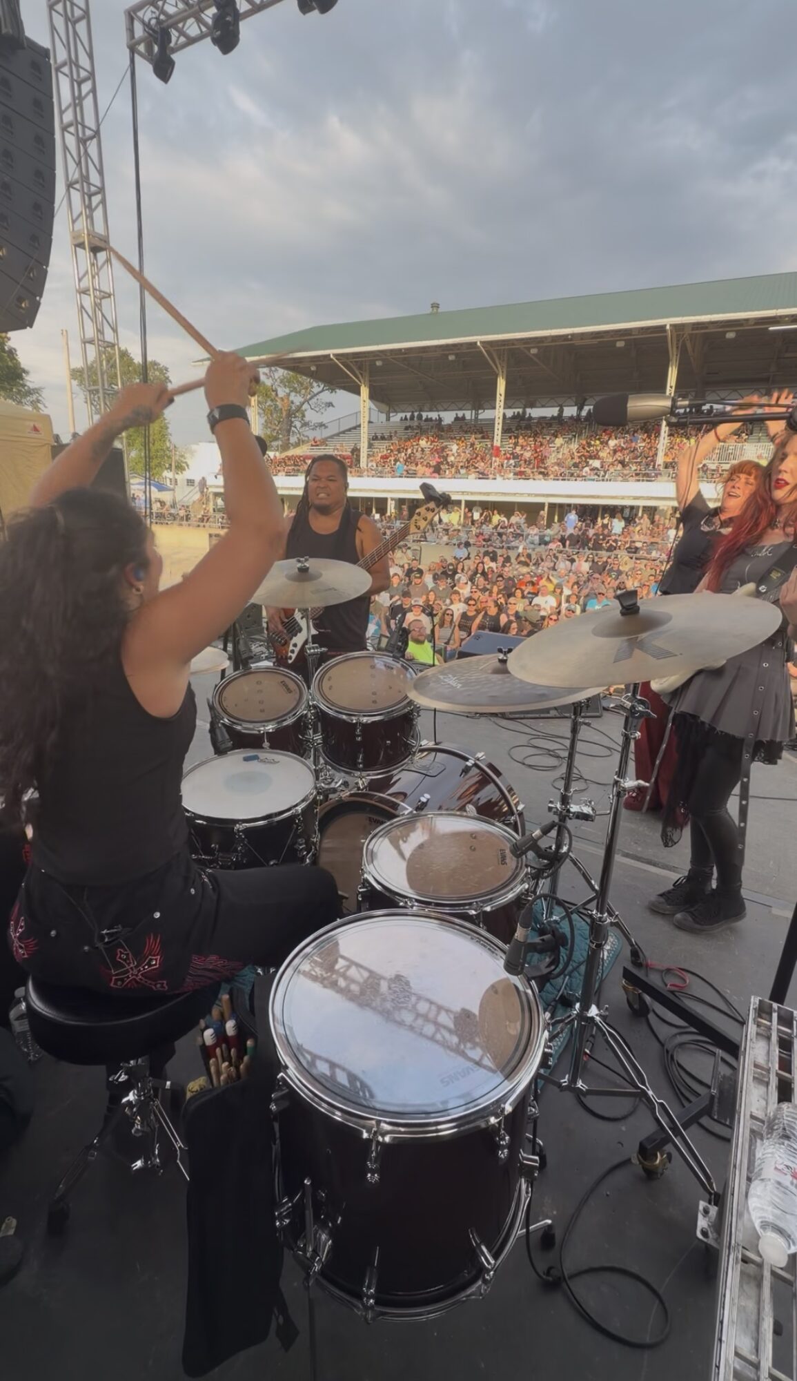 A drummer playing on stage with a large crowd in the background, drums and cymbals visible, outdoor concert setting.