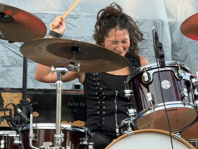 Woman playing drums, smiling, with drumsticks, surrounded by drums and cymbals, in a performance setting.