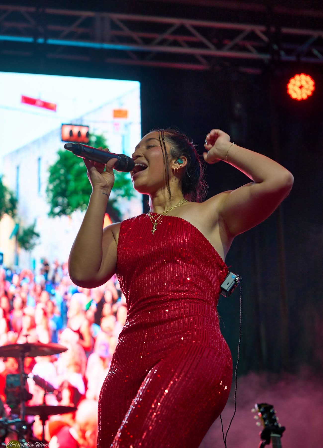 Woman singing into microphone on stage, wearing red dress, with crowd and stage lights in background.