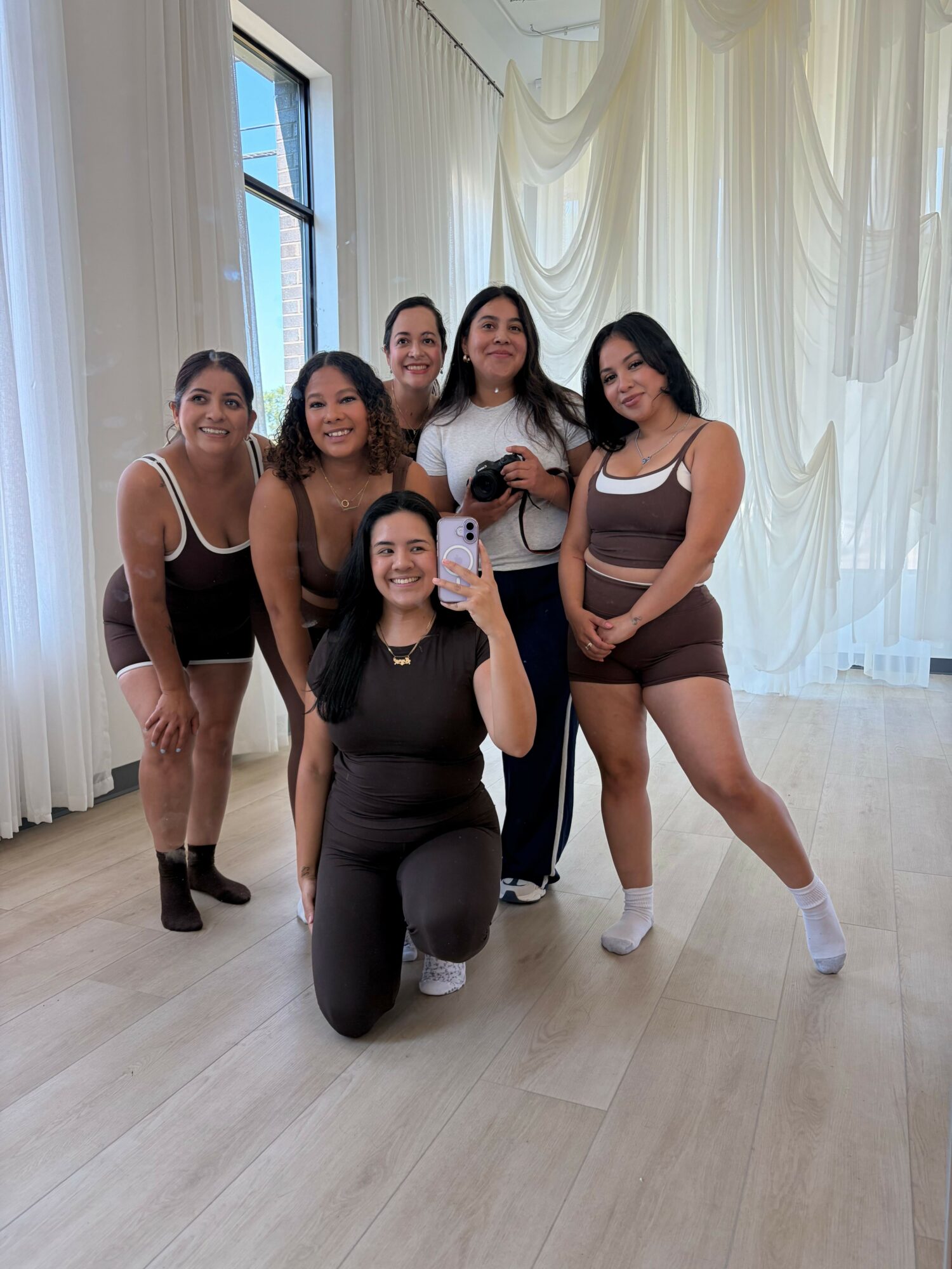 Five women posing together indoors, some standing and one kneeling, with light-colored curtains and a window in the background.