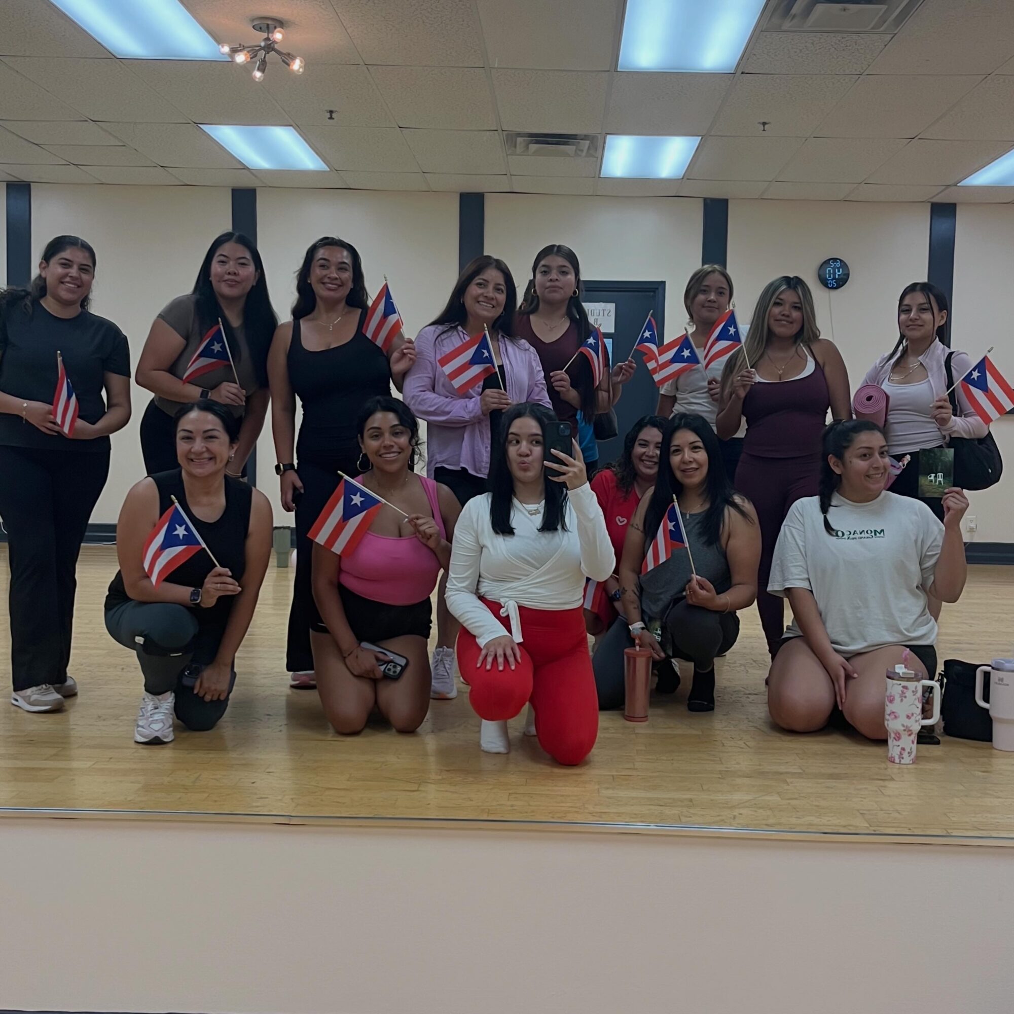 Group of women holding Puerto Rican flags, posing indoors on a wooden floor, some kneeling and some standing.