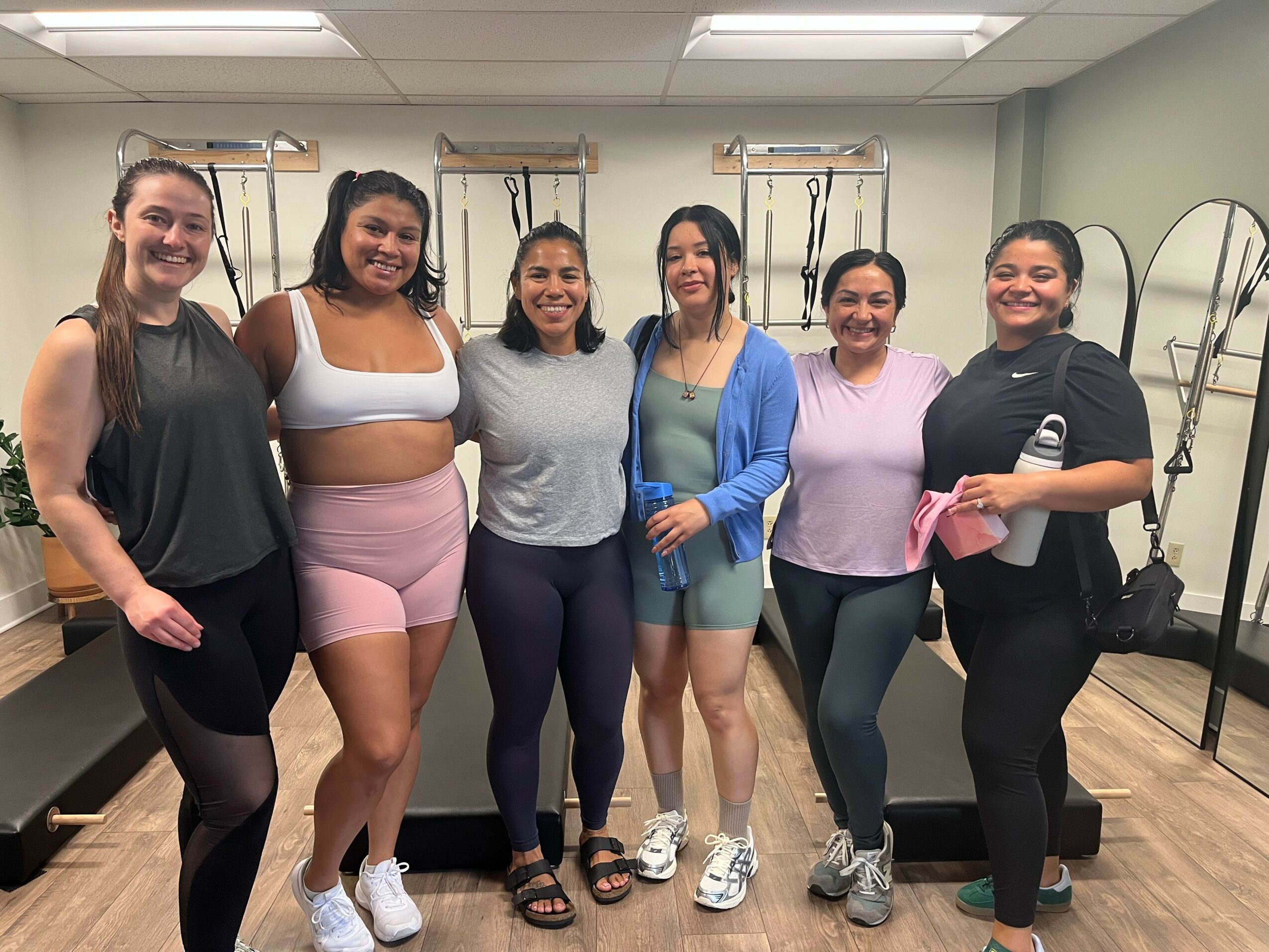 Six women standing together in a room with mirrors and exercise equipment, smiling at the camera.