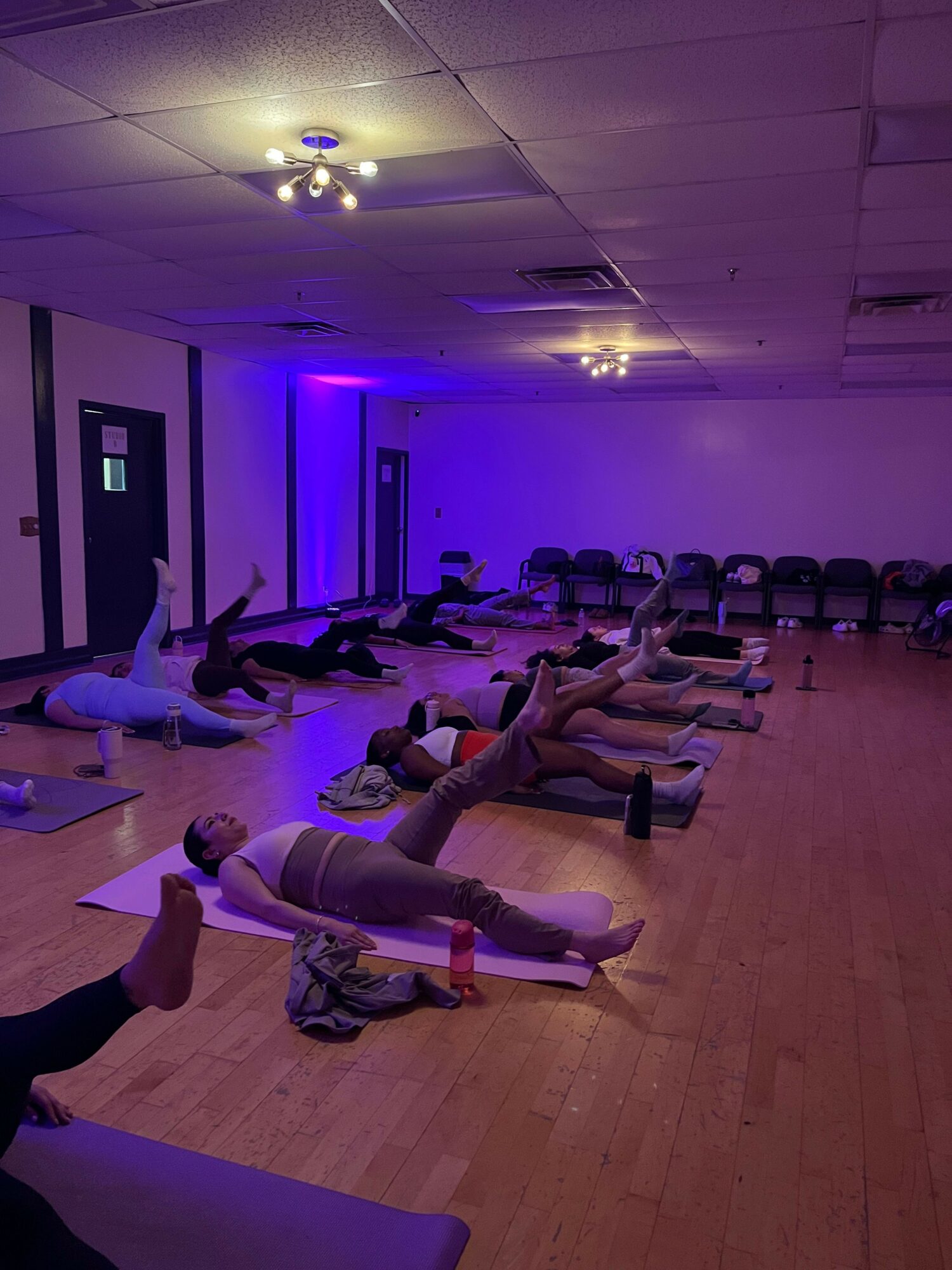 Group of people practicing yoga on mats in a room with purple lighting, wooden floor, and white ceiling.