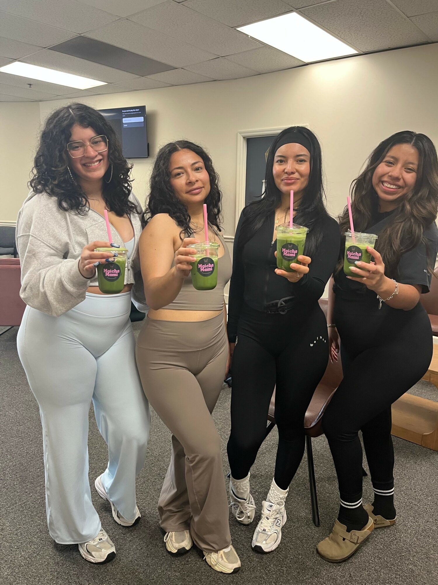 Five women in a mirror selfie holding drinks, smiling, in a room with ceiling lights and a door in the background.