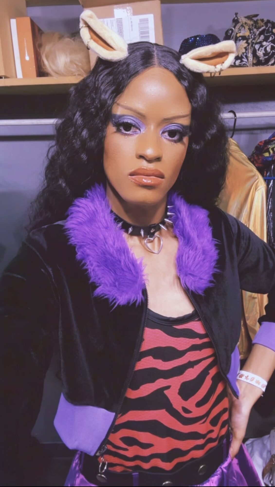Woman with curly hair wearing bunny ears, purple fur collar, zebra print top, and choker, posing indoors.