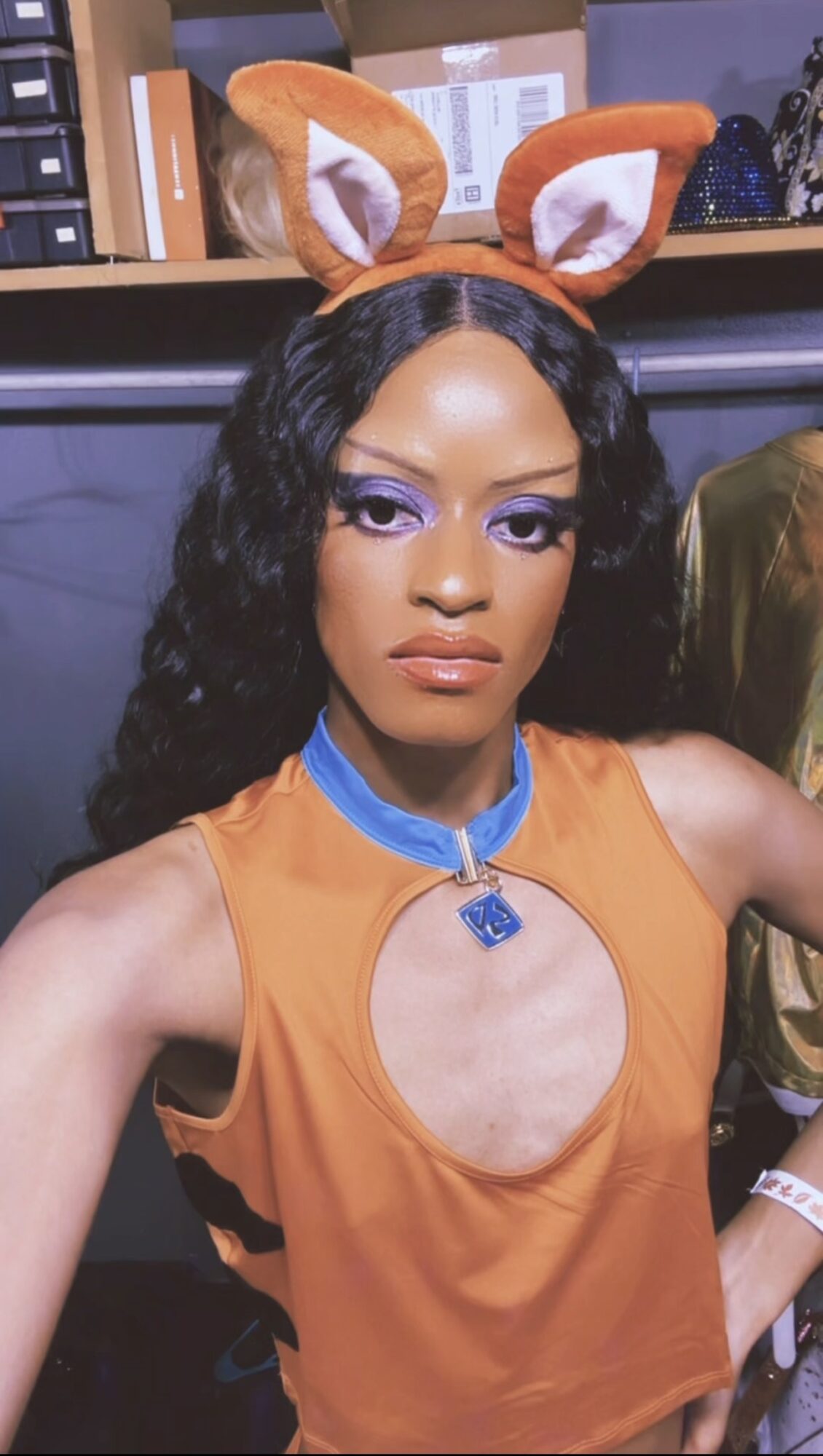 Person wearing a fox costume with orange ears, purple eyeshadow, and a sleeveless orange top, standing indoors.