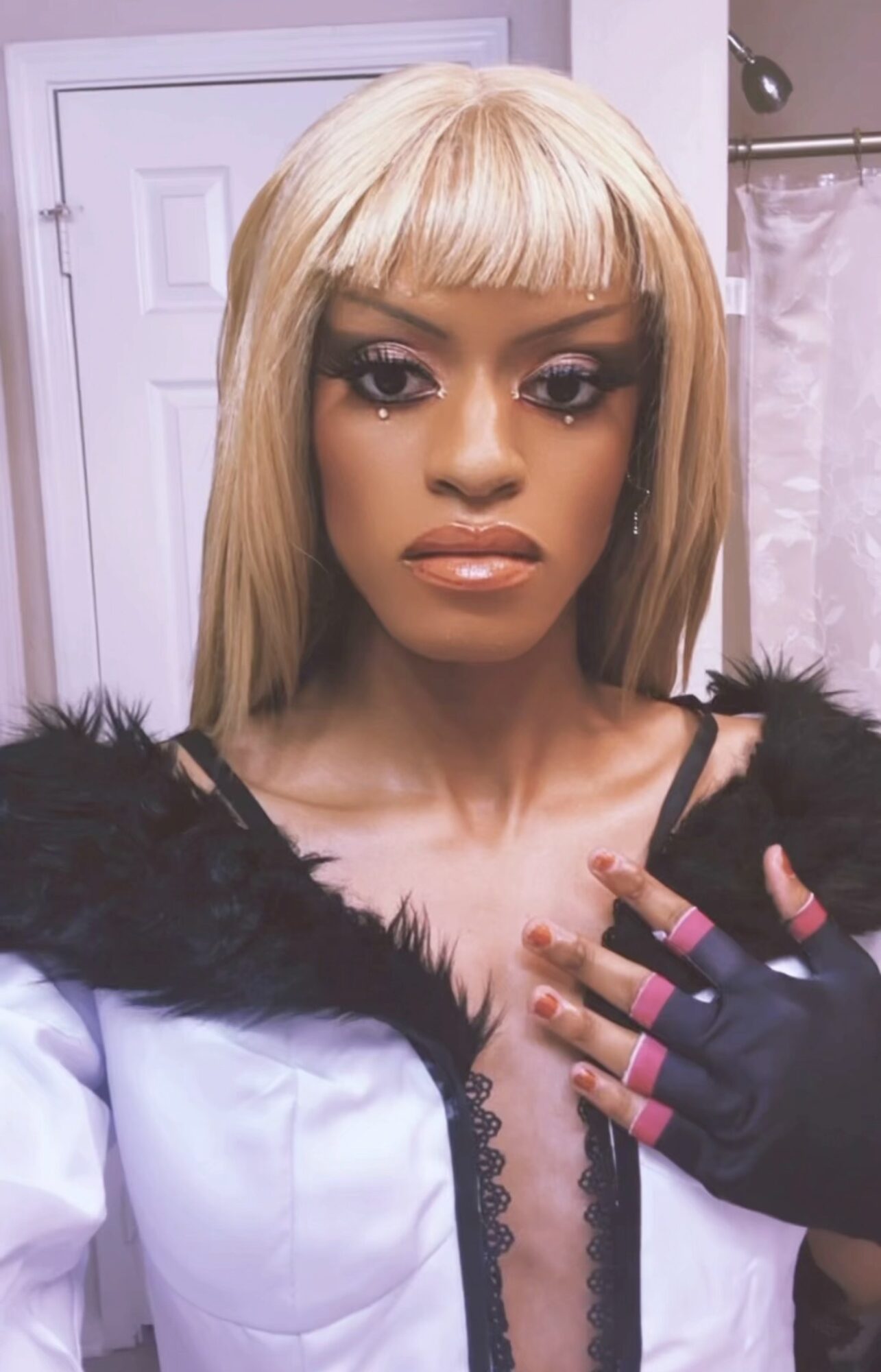 Woman with blonde hair and makeup, wearing a white top with black fur trim, in a bathroom.