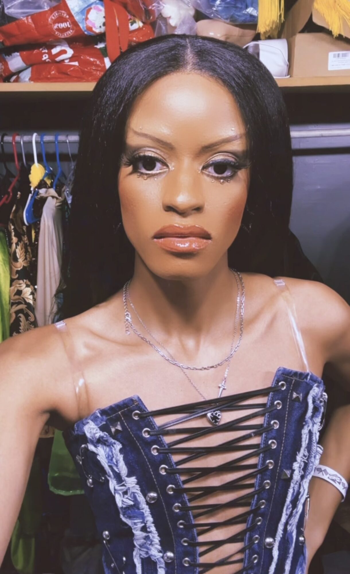 Woman with long black hair and makeup, wearing a blue lace-up top, in a room with clothing and shelves