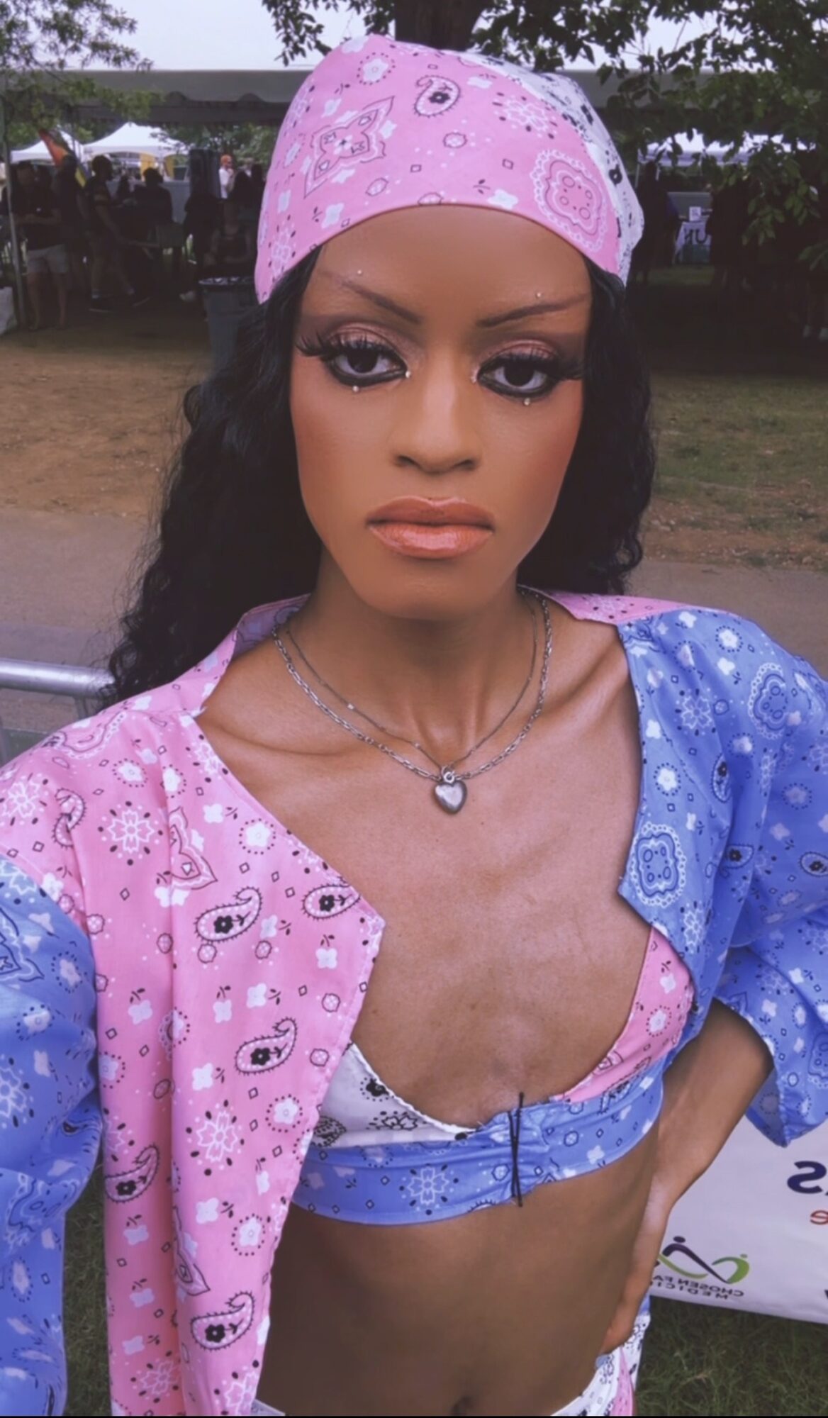 Young woman with dark hair, wearing a pink bandana and a blue and pink patterned top outdoors.