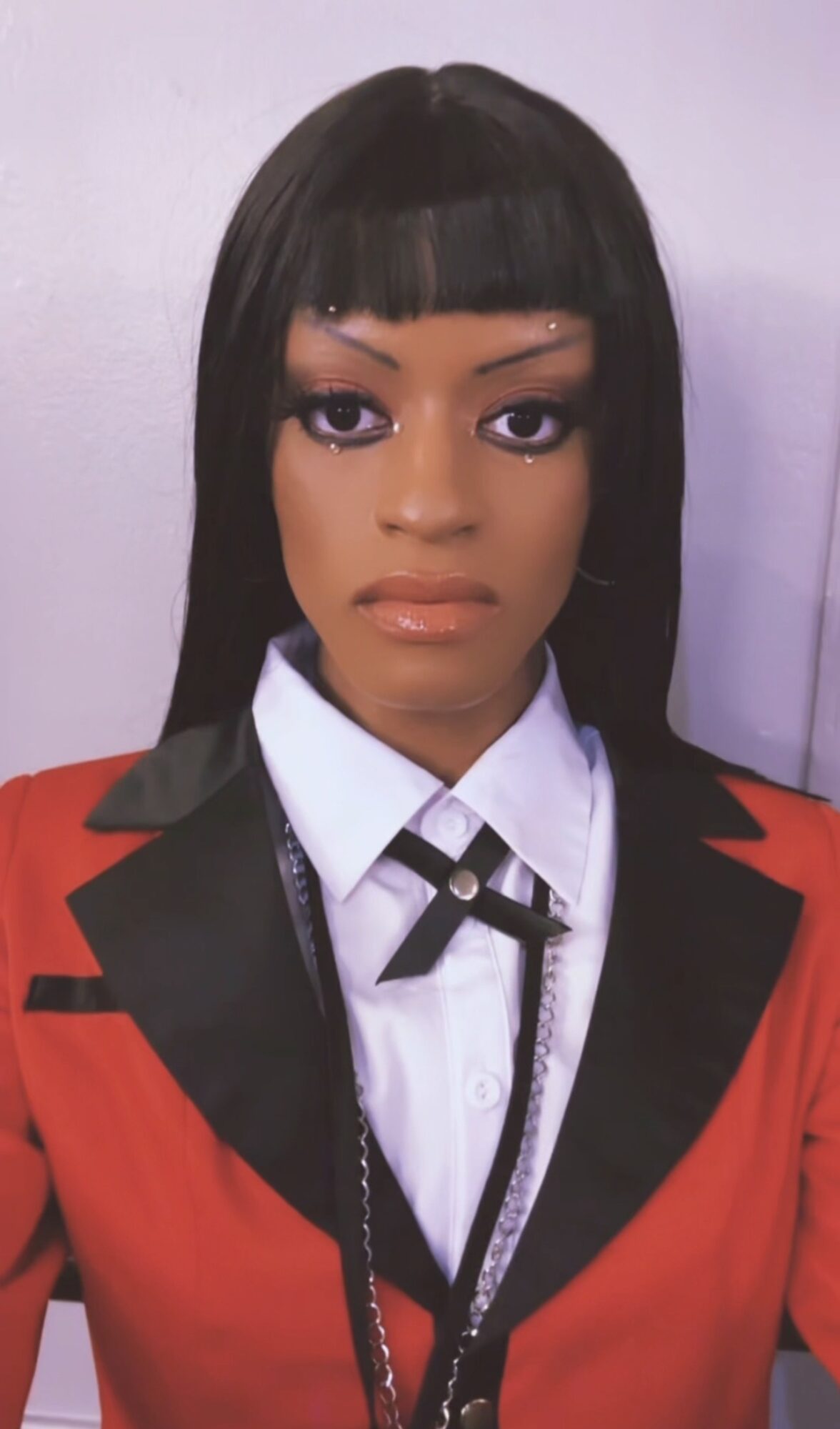 Woman with straight black hair and bangs, wearing a red blazer with black lapels, white shirt, and black tie, against a plain background.