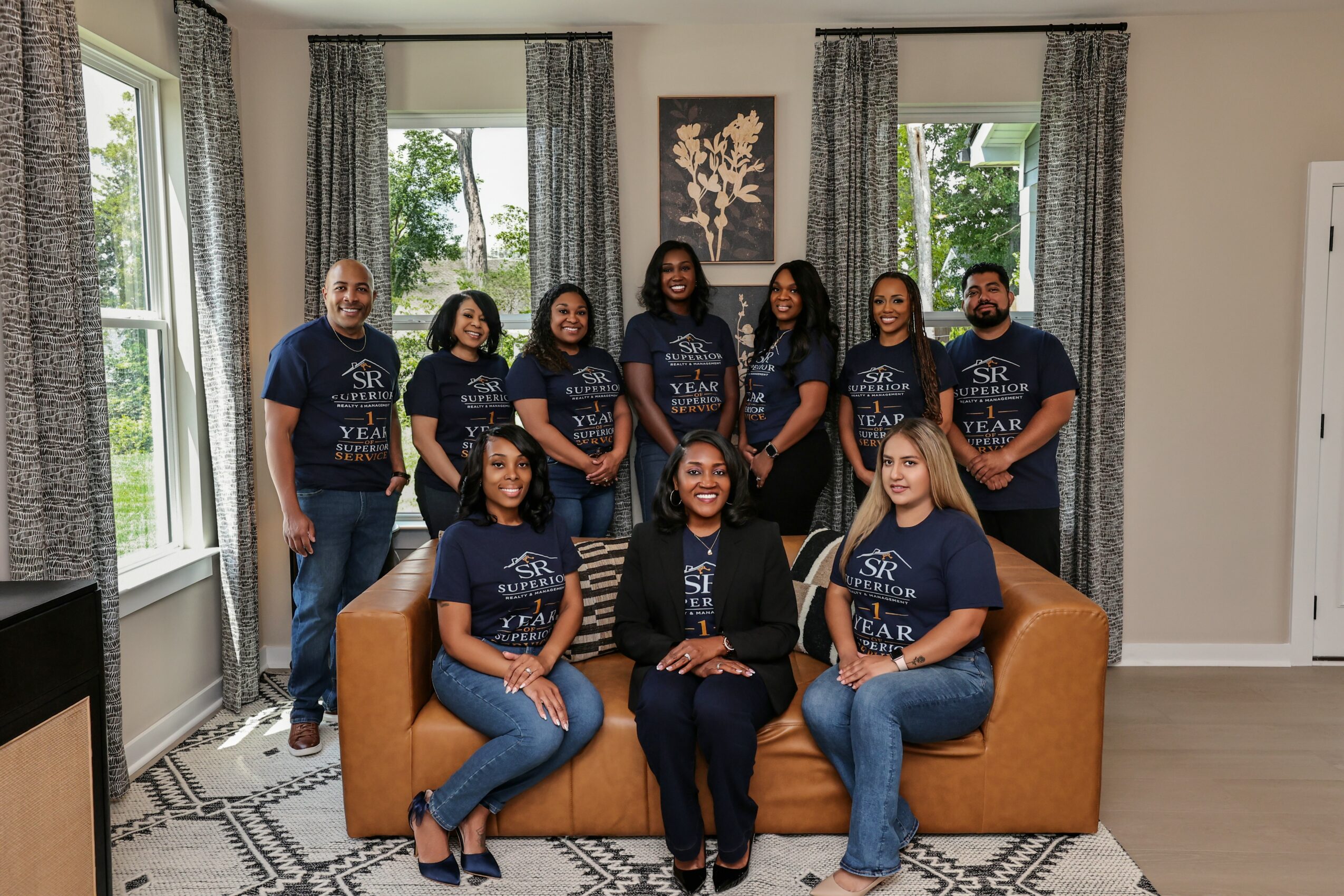 Group of nine people in matching navy T-shirts sitting and standing in a living room with large windows and curtains.
