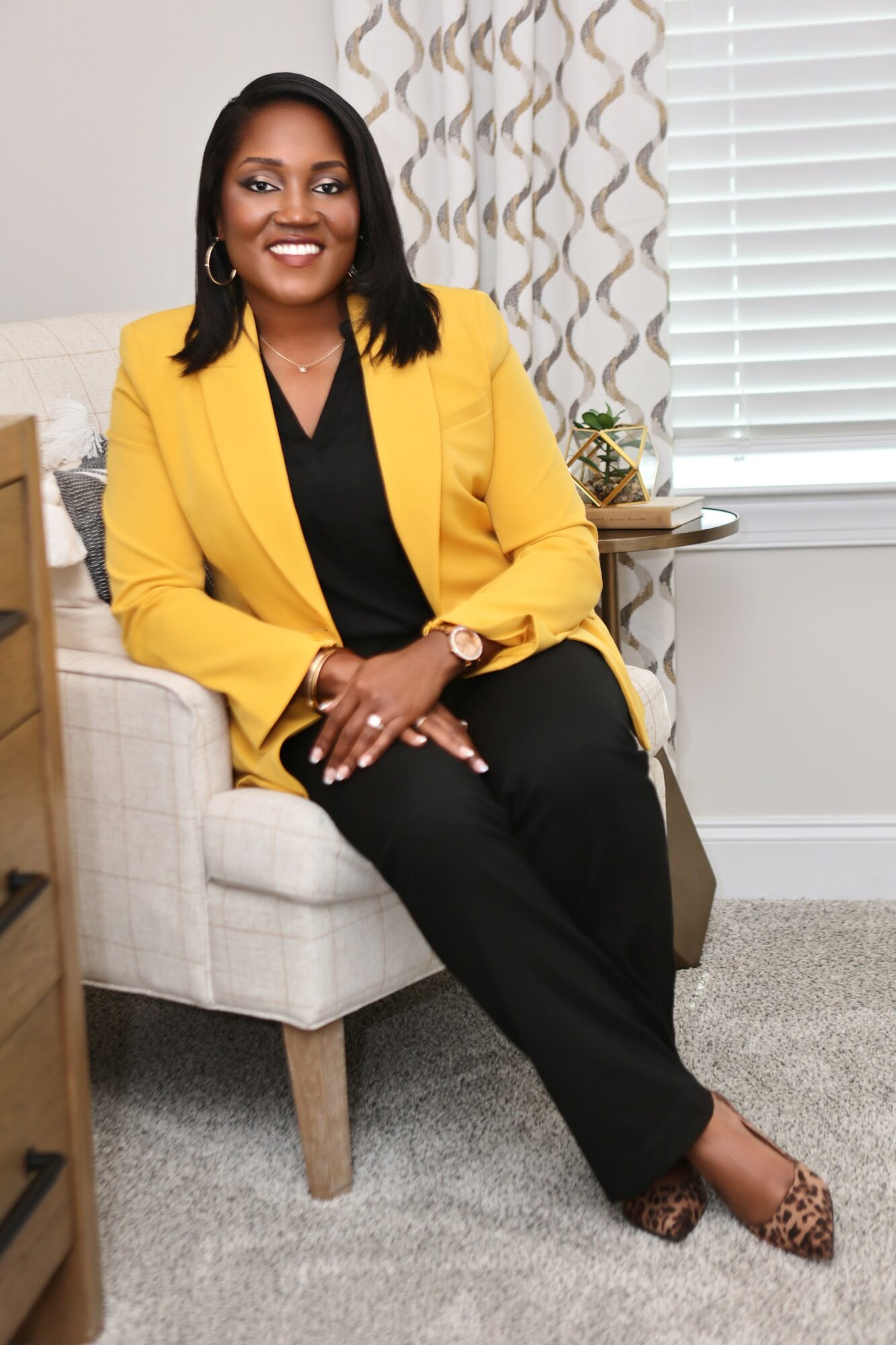 Woman sitting on a chair, smiling, wearing a yellow blazer and black pants, in a room with window blinds and a side table.