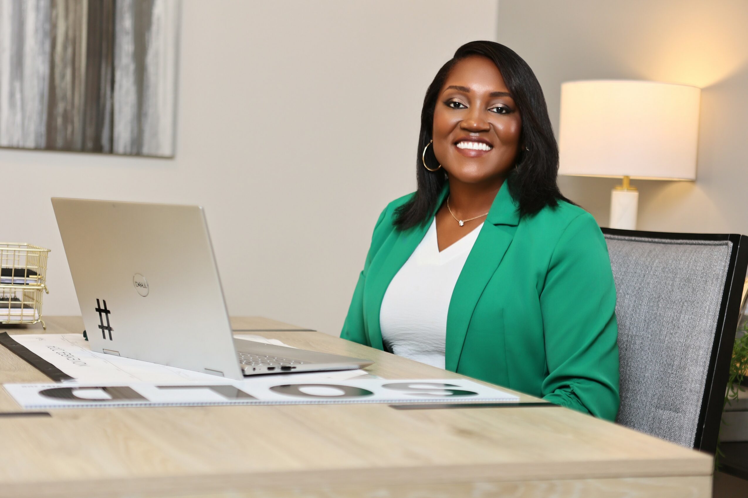 Smiling woman with dark hair, wearing a green blazer, sitting at a desk with a laptop and papers, in an office setting.
