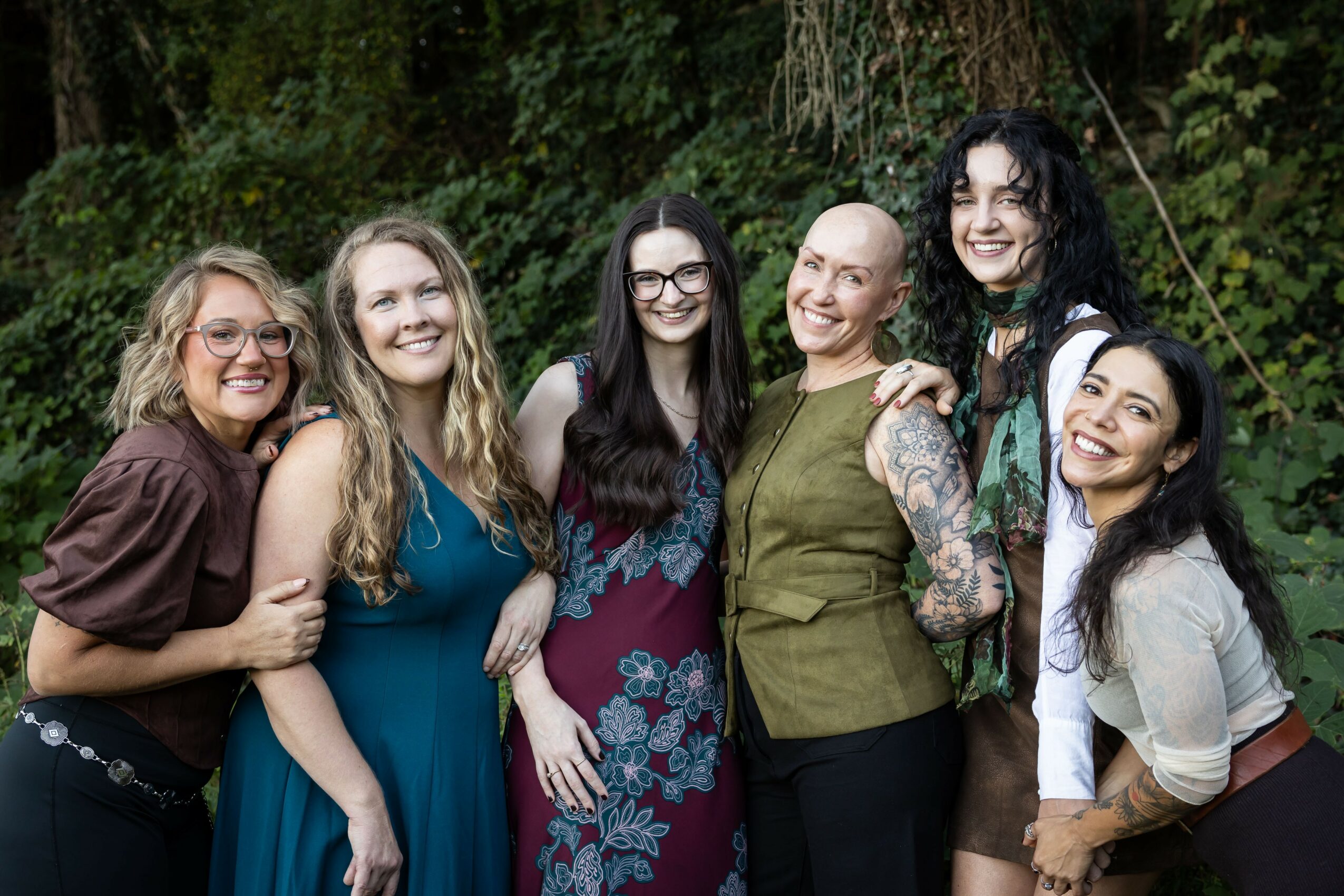 Six women standing outdoors in front of greenery, smiling and posing together, some with arms around each other.