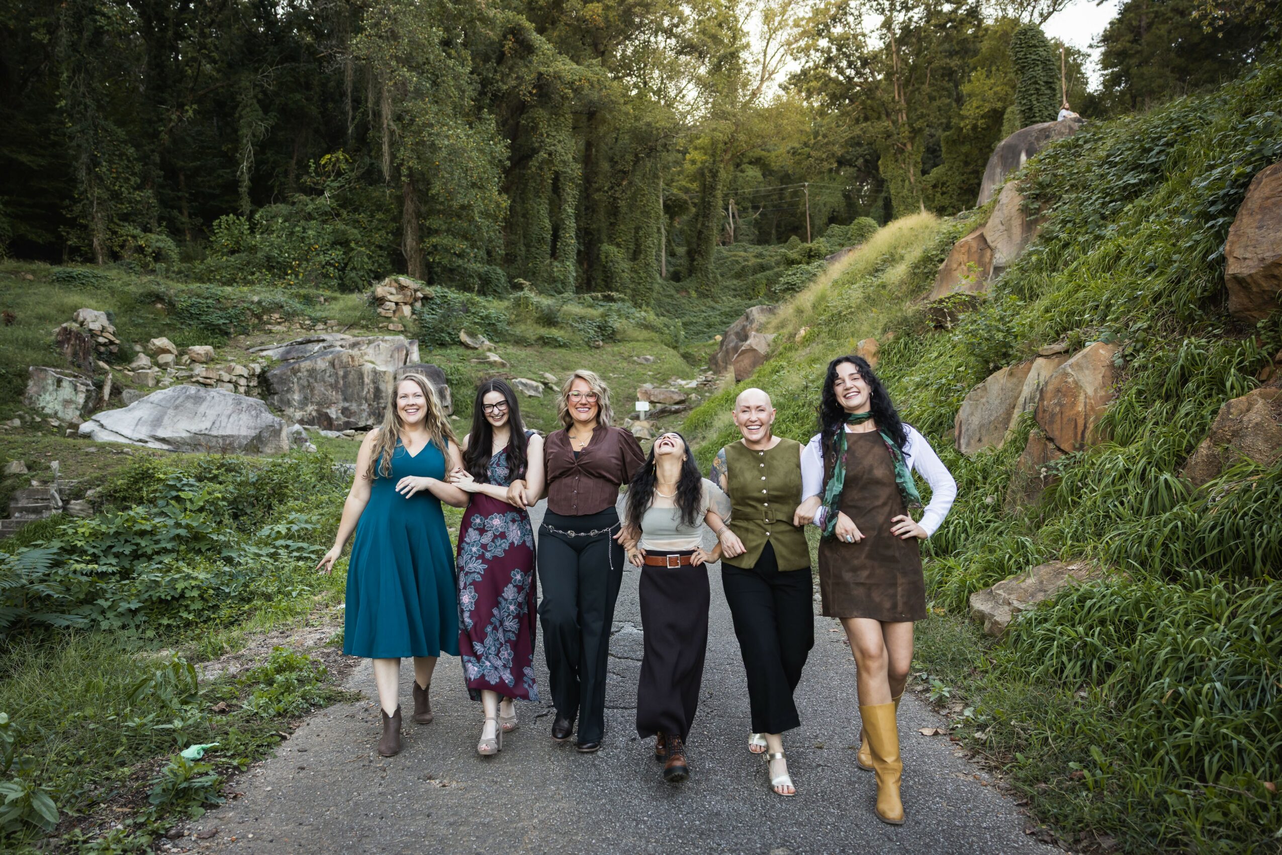 Five women walking on a path in a lush, green park with rocks and trees in the background.
