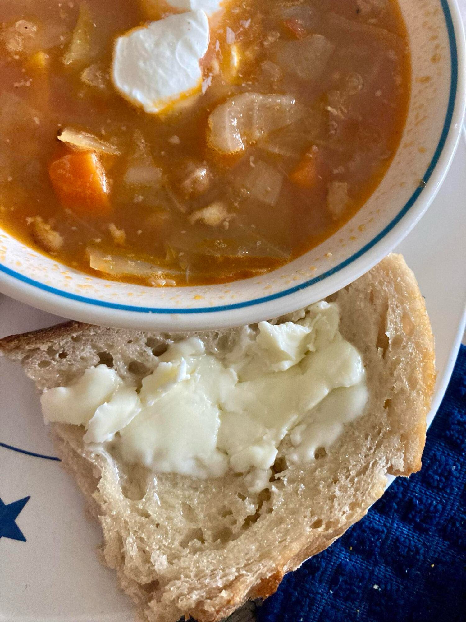 Bowl of vegetable soup with carrots, potatoes, and a dollop of sour cream, served with a bread slice topped with butter or cheese.