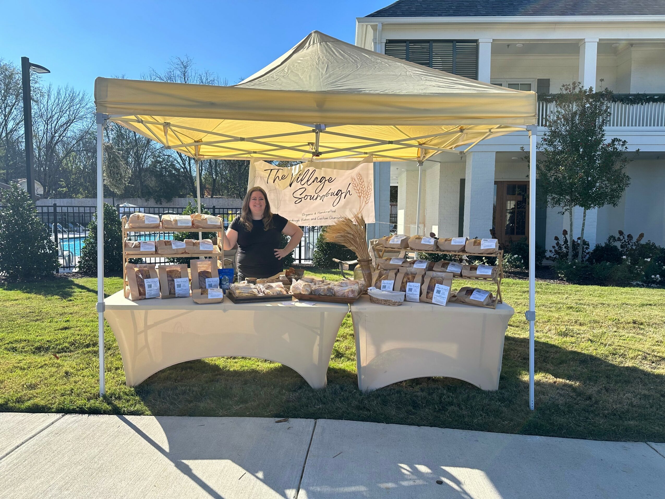 Person standing behind a table with baked goods under a yellow canopy outdoors, with a house and trees in the background.