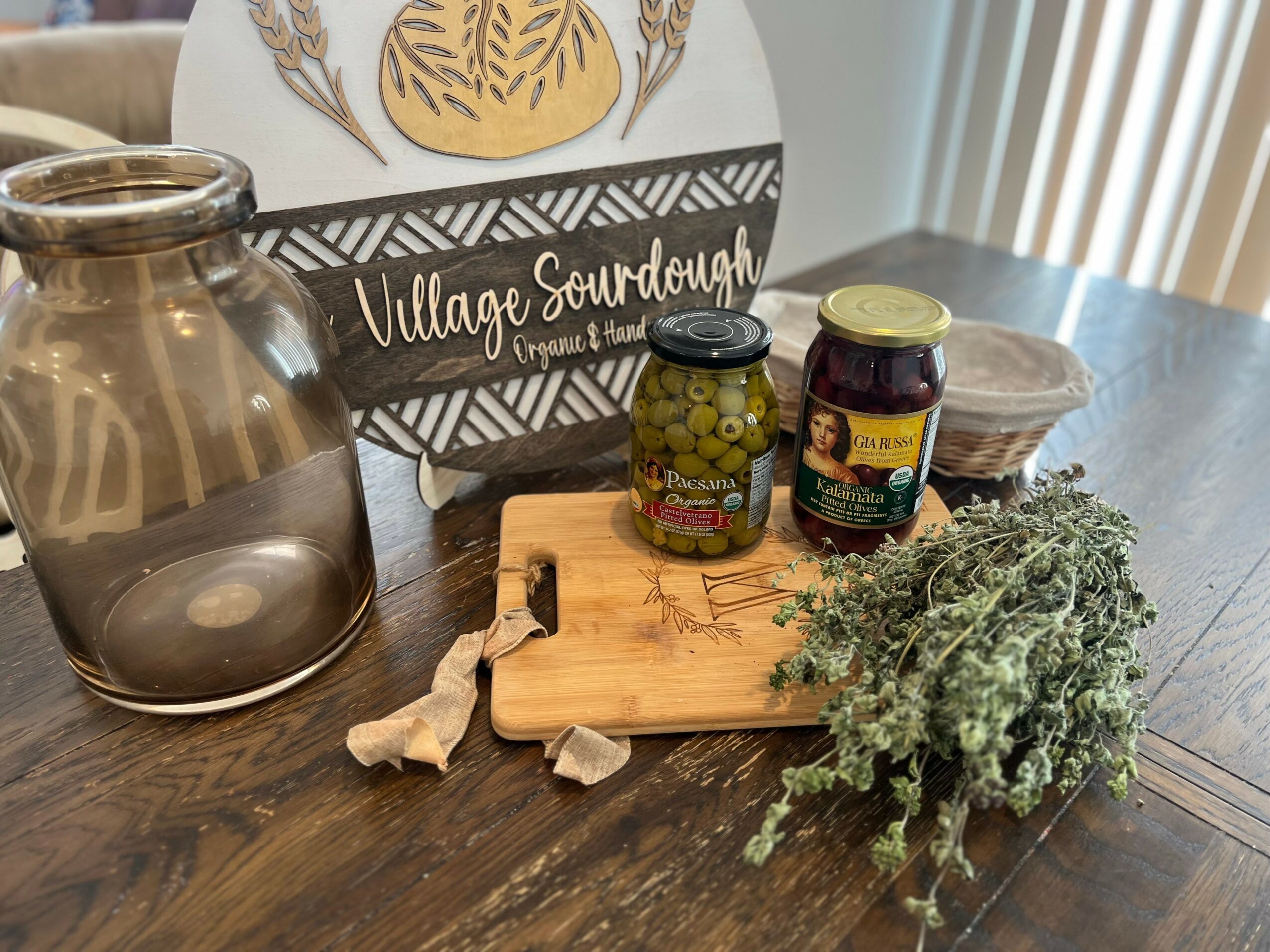 Jar, wooden cutting board with jars of jam, dried herbs, and a bunch of fresh herbs on a wooden table.