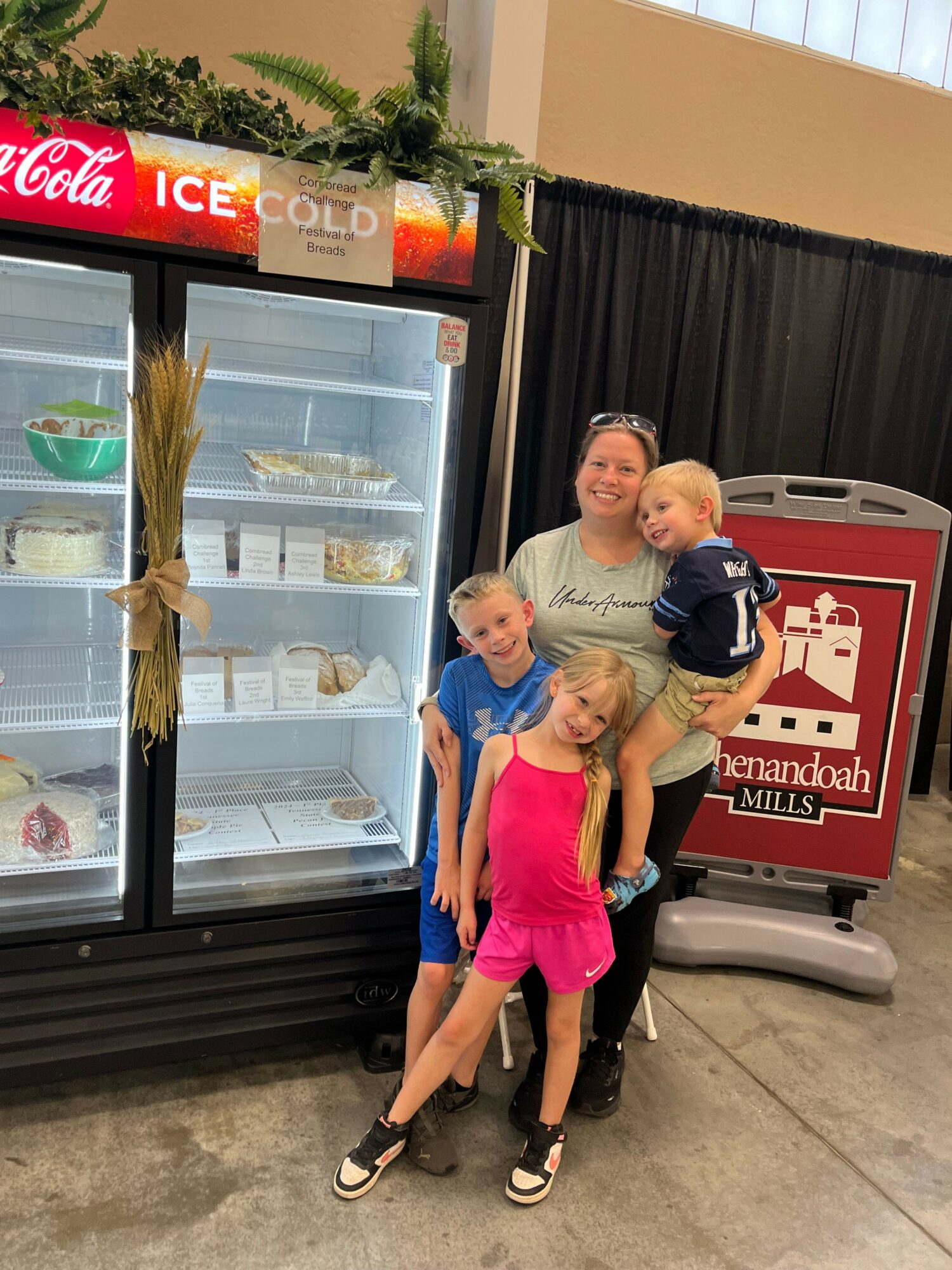 Three people, two children and an adult, stand in front of a refrigerator with glass doors, in an indoor setting. The children are a girl in pink and a boy in blue, and the adult is a woman. They are smiling and posing for the photo.