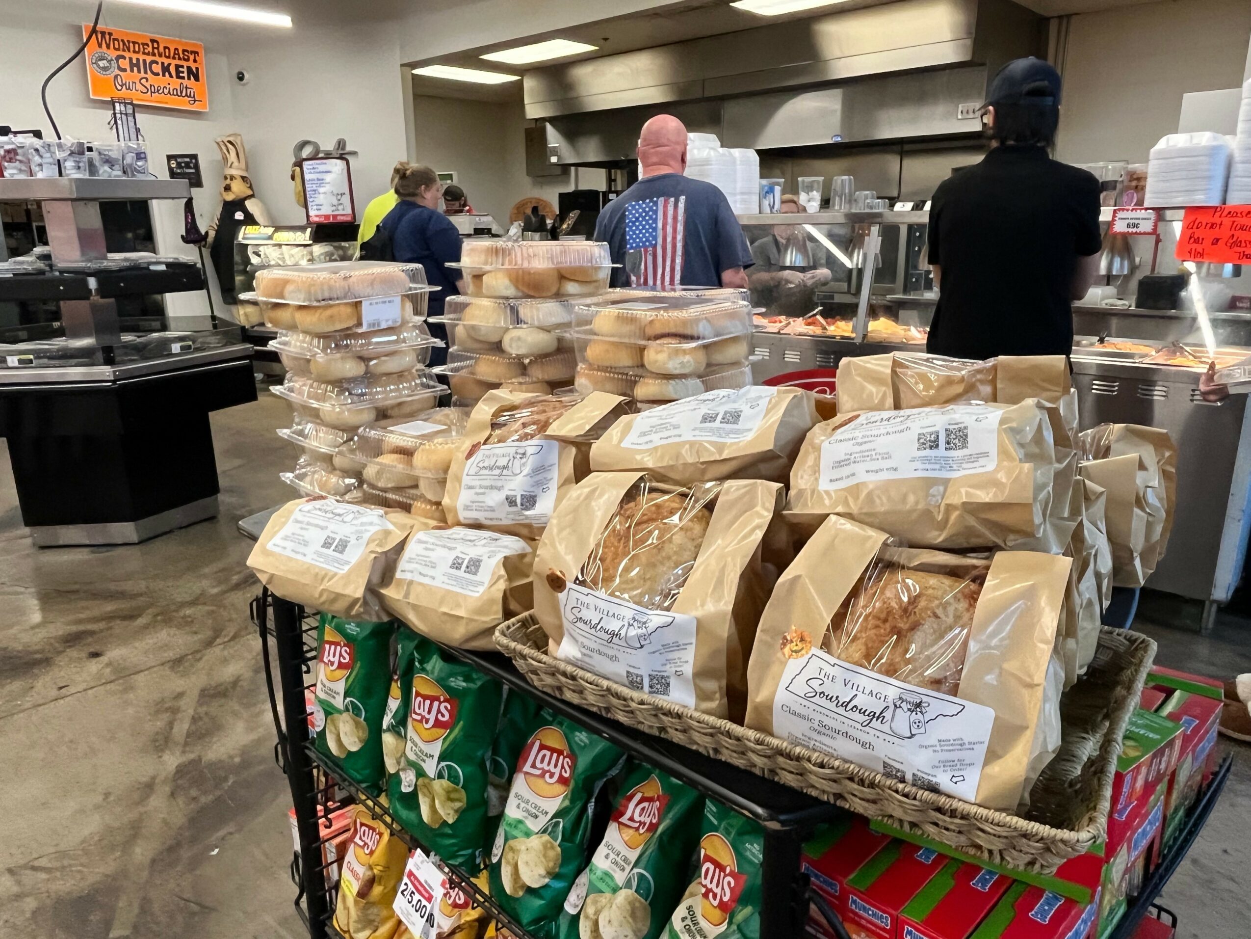 Bags of bread and packs of Lay's chips on a display in a store, with people working behind the counter.
