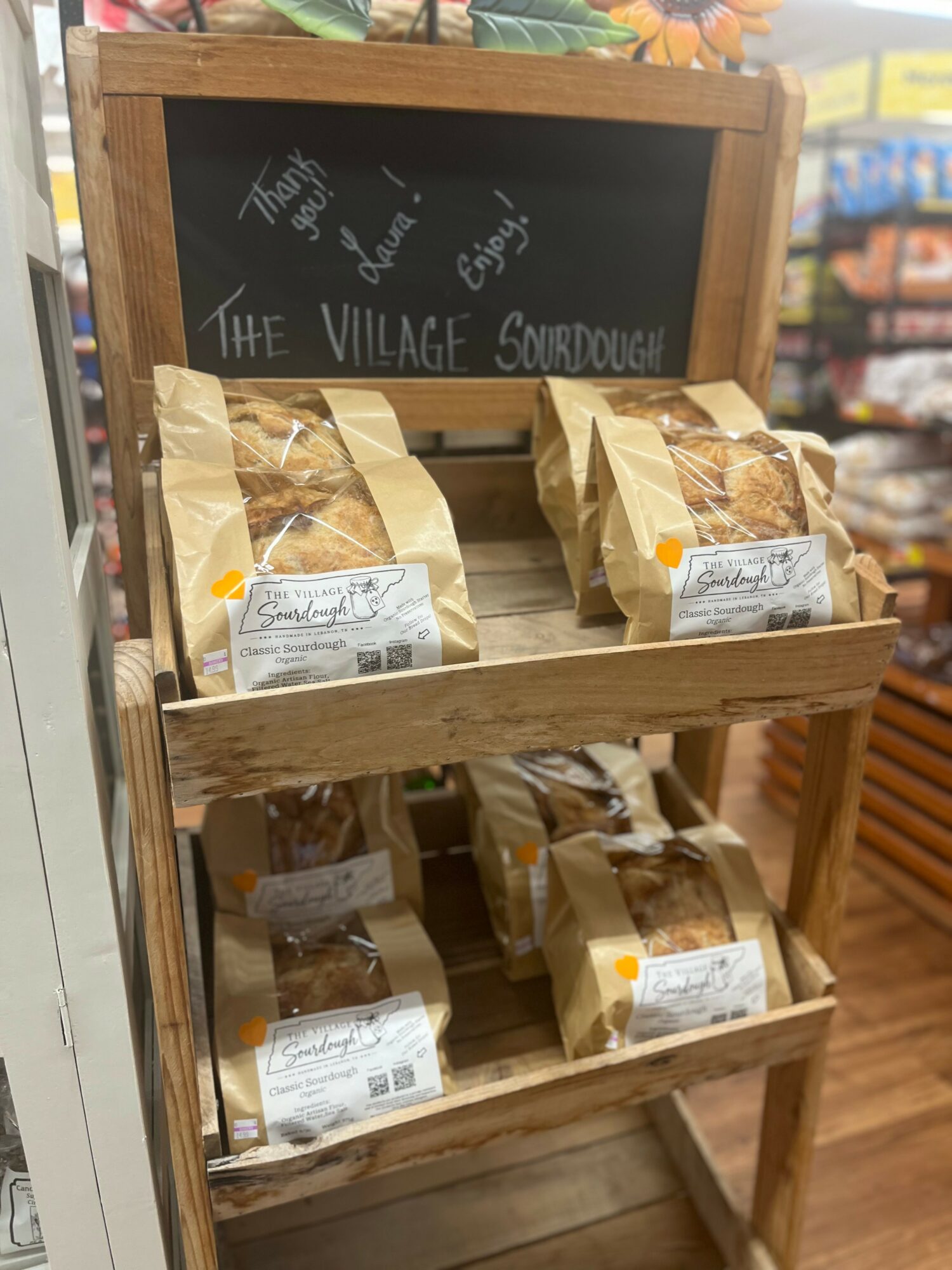 Wooden display stand with bags of baked goods on two shelves, chalkboard sign on top, in a store setting.
