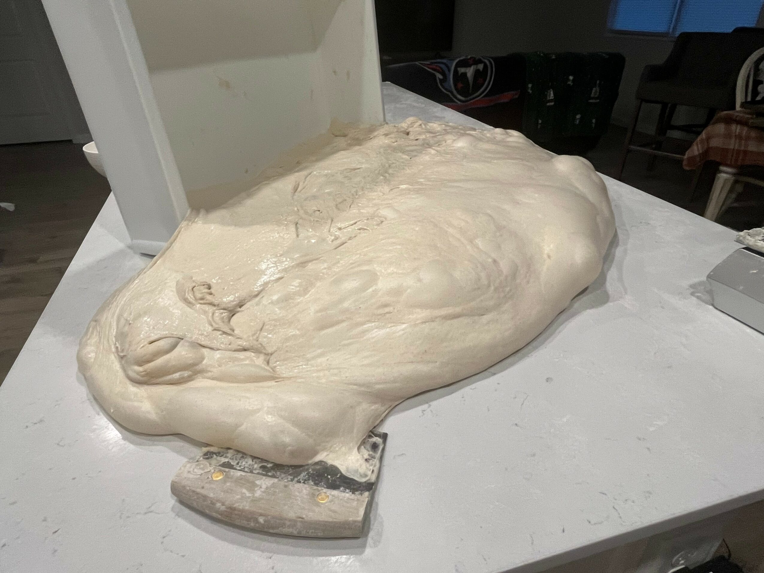 Dough resting on a white surface, partially covered by a white sheet, in a kitchen or bakery setting.