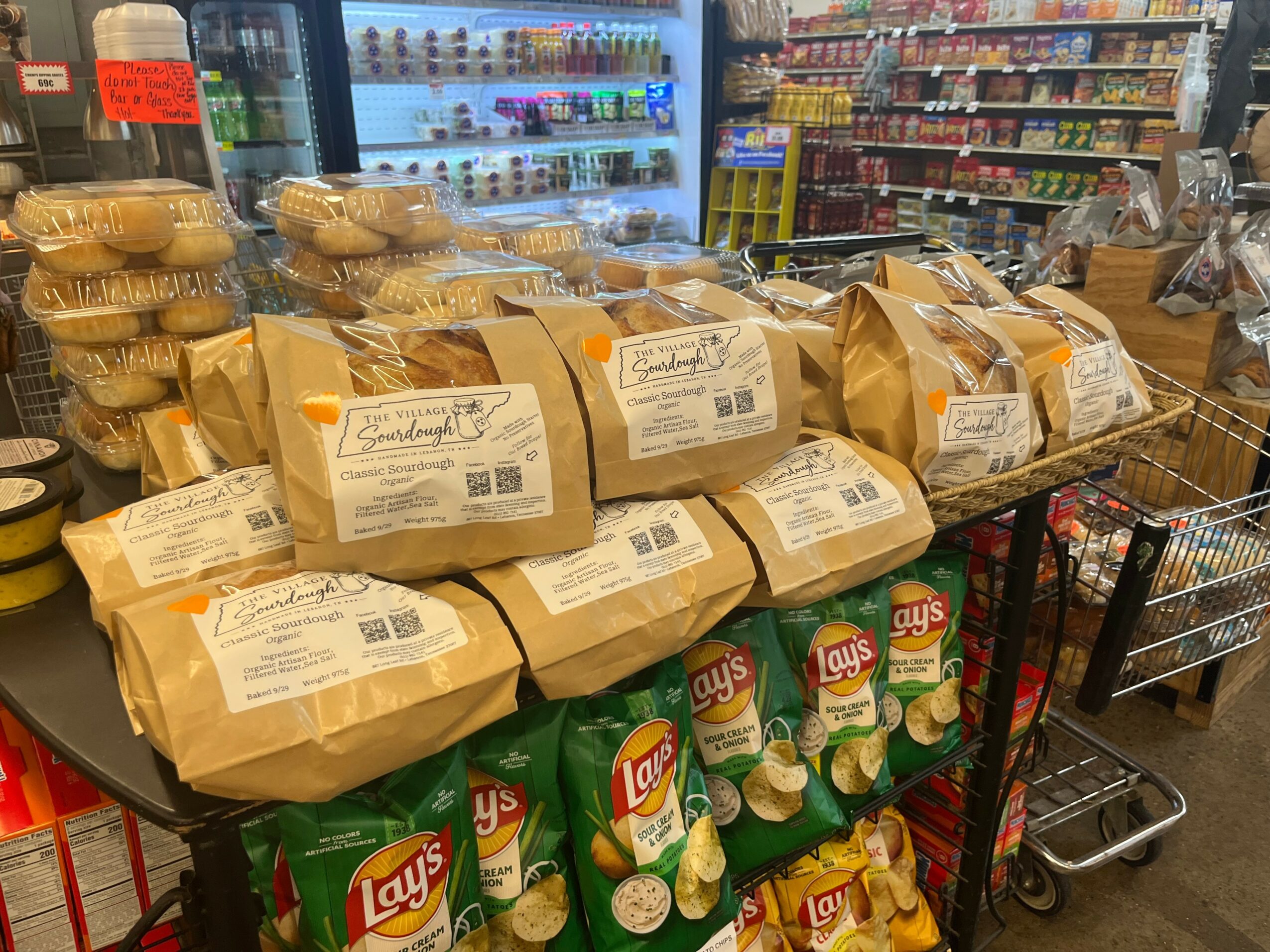 Display of snack bags and baked goods on a shopping cart in a grocery store, with shelves and refrigerated items in the background.