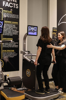 Two women in black clothing using a medical or fitness device in a room with informational posters on the wall.