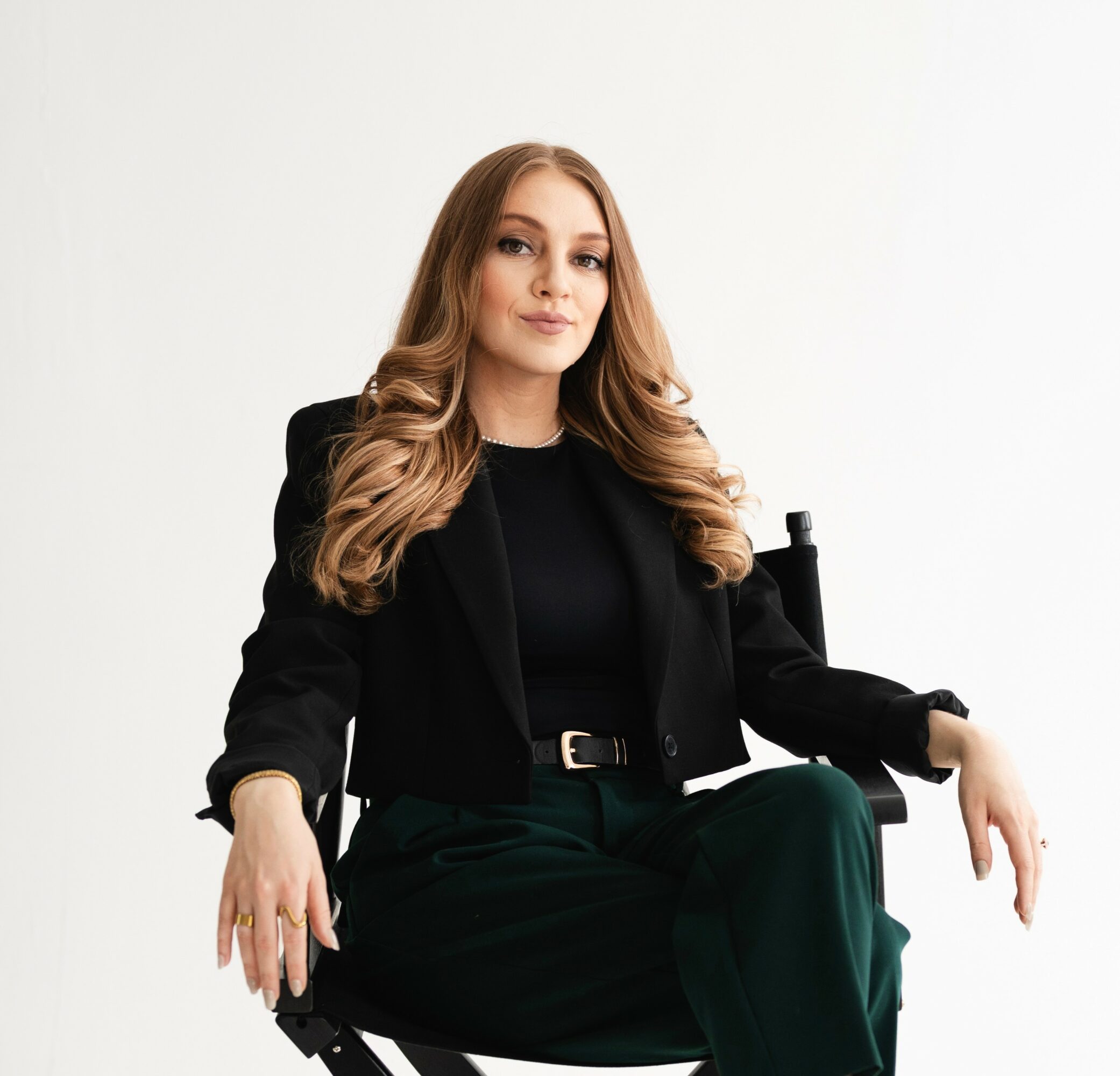 Woman with long wavy hair sitting in a chair, wearing a black blazer and dark green pants, against a white background.