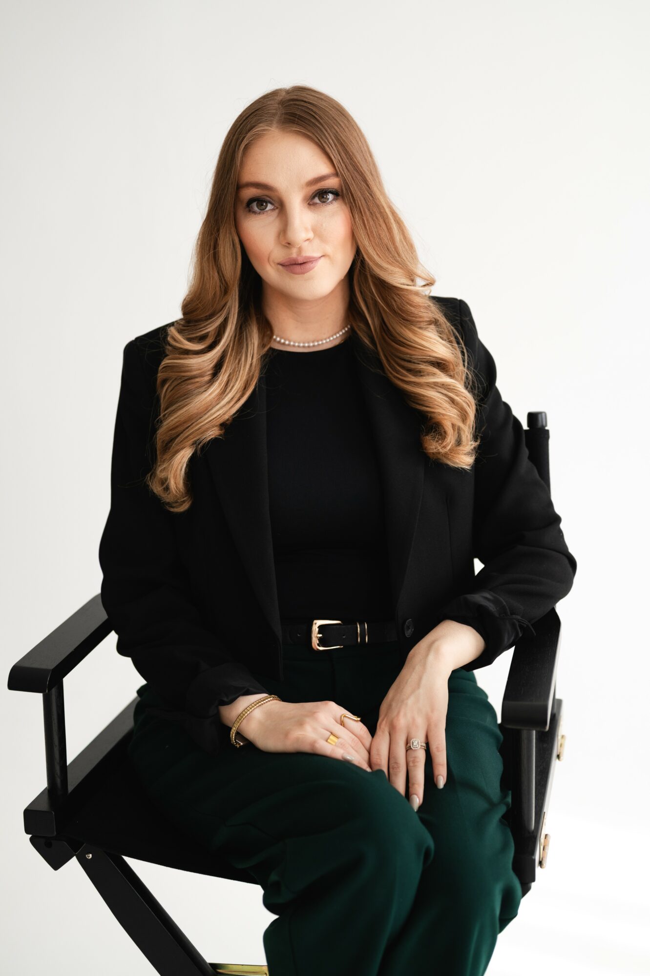Young woman with long wavy hair sitting in a black chair, wearing a black blazer and dark green pants, smiling softly.