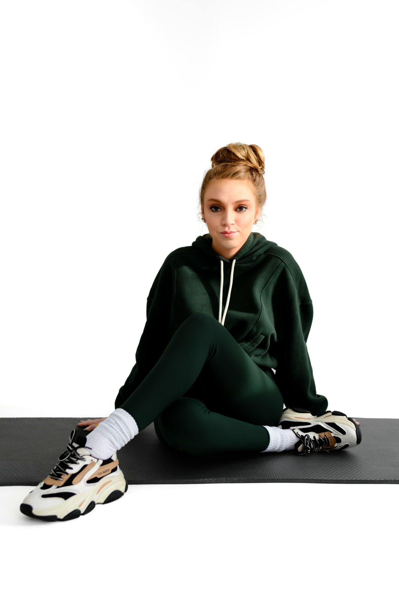 Young woman with red hair in a bun, wearing a dark green hoodie, black leggings, and sneakers, sitting on the floor against a white background.