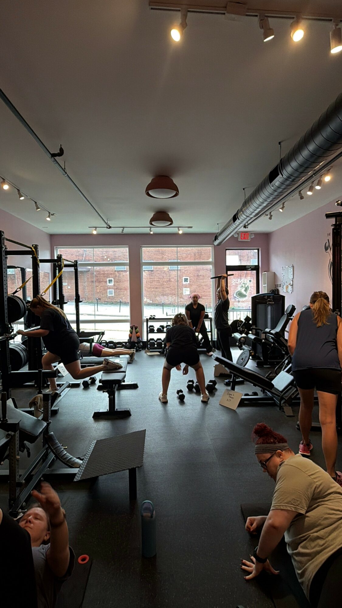 People exercising in a gym with large windows, workout equipment, and ceiling lights, some seated and others standing or stretching.