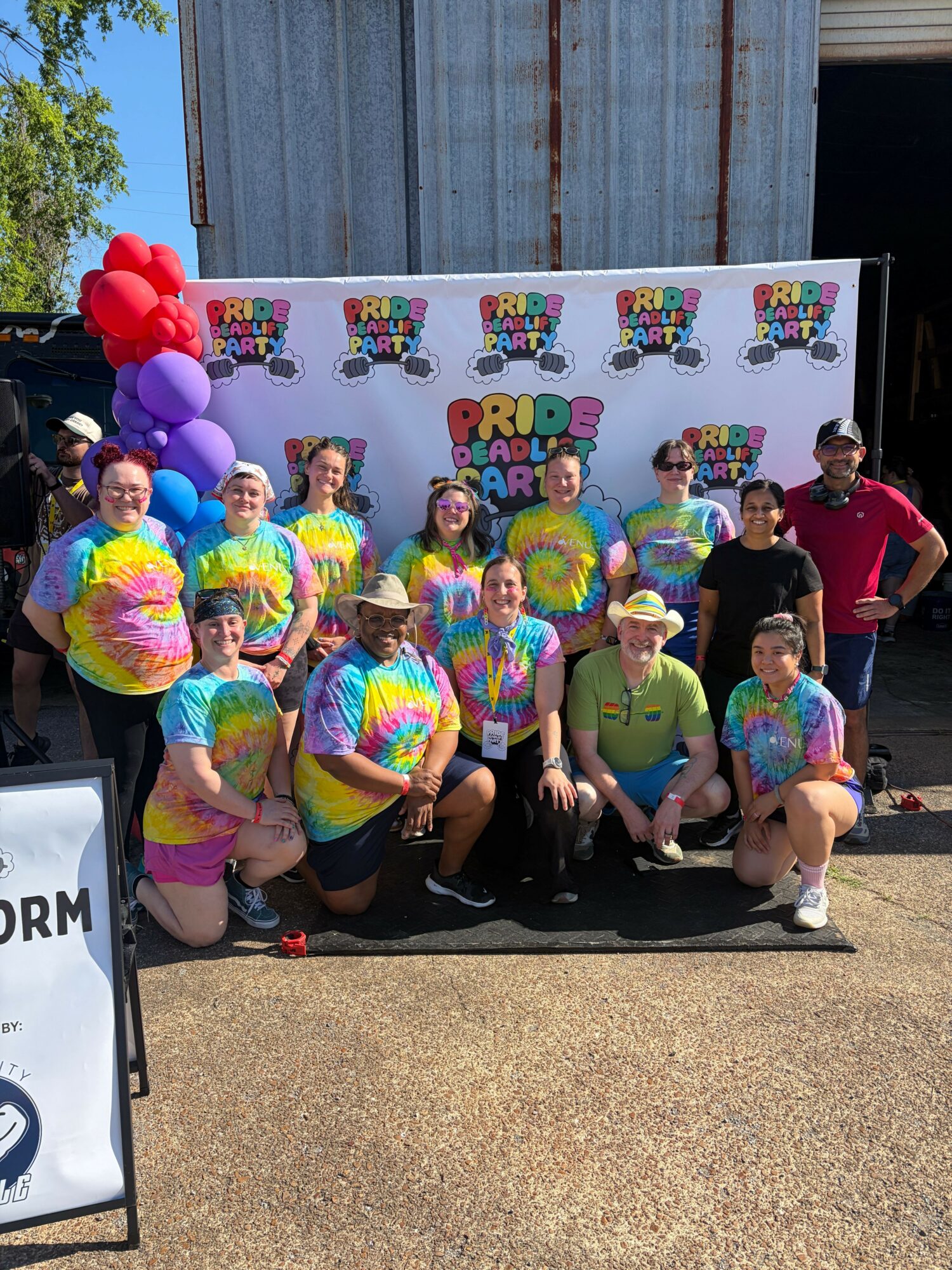 Group of people in colorful tie-dye outfits posing outdoors with a decorated backdrop and balloons.