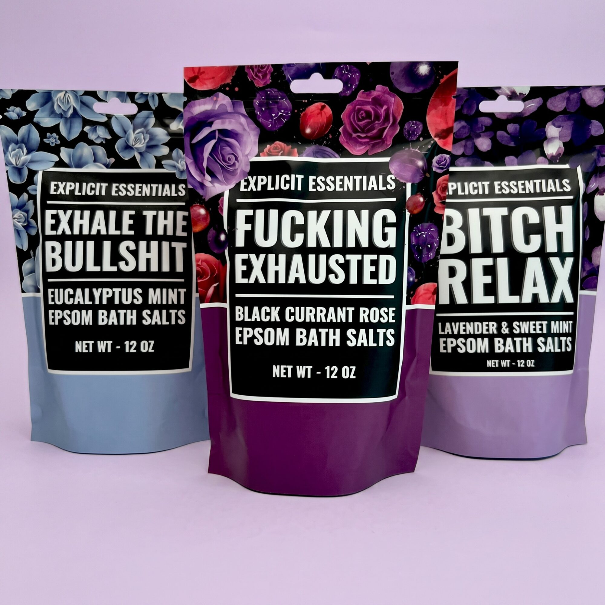 Three bath salt packages with humorous labels, featuring purple, black, and lavender colors, and floral designs on top. Each package has a different phrase.