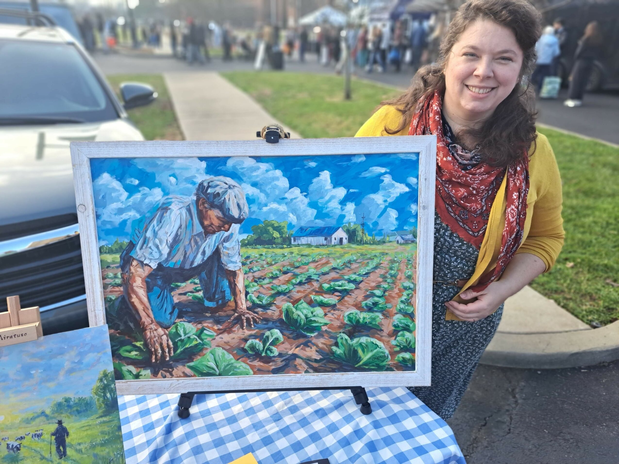 Woman standing outdoors next to a framed painting of a person working in a garden, with a crowd and cars in the background.