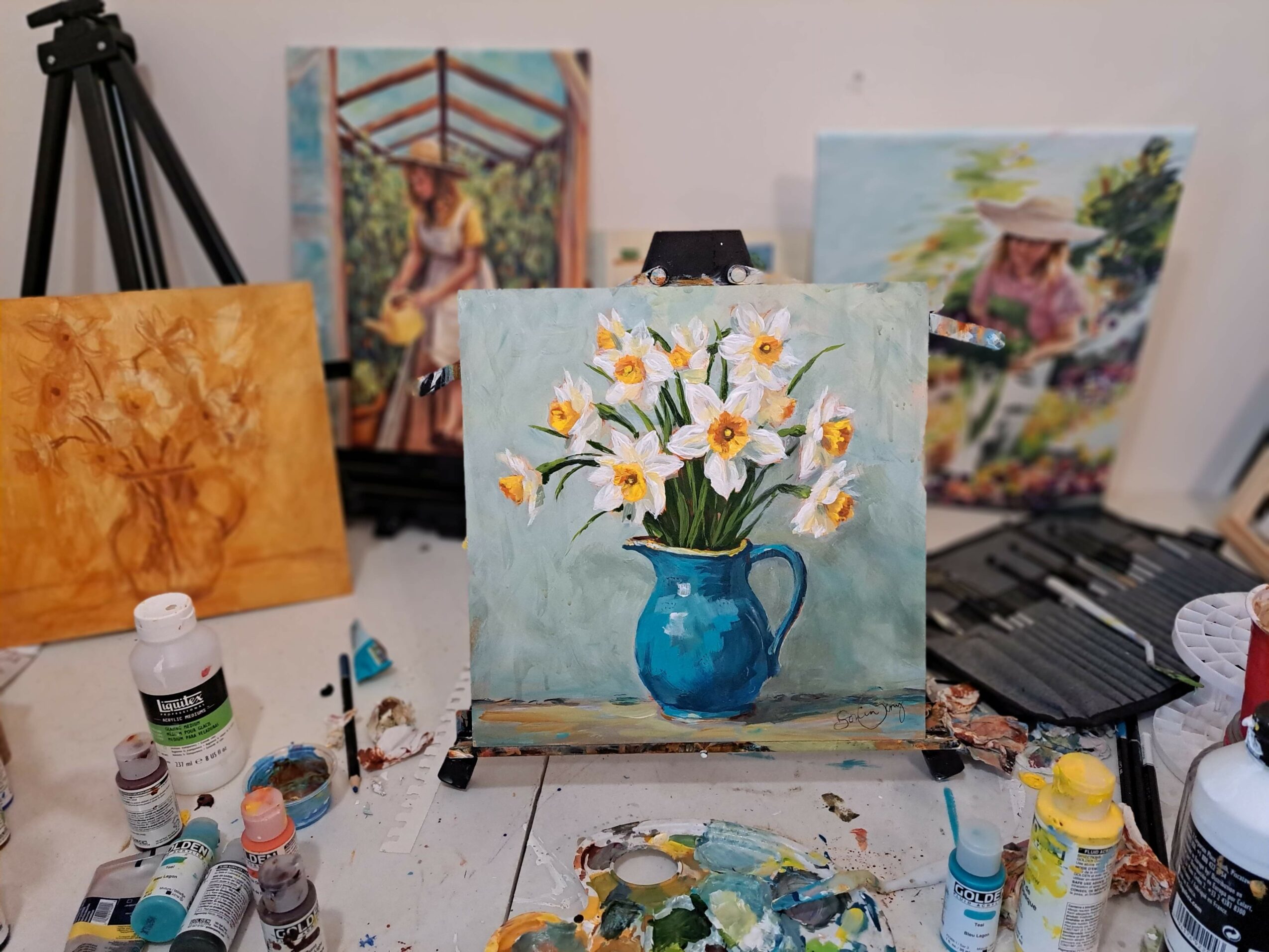 Small painting of white flowers in a blue vase on a table, with art supplies and other paintings in the background.