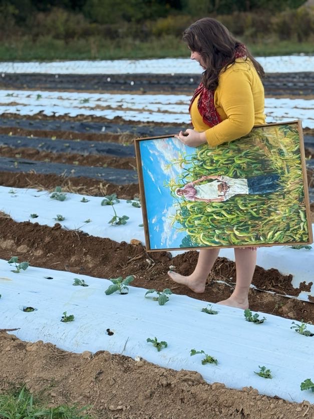 Woman standing barefoot in a field holding a painting of a woman in a white dress with a pond background.