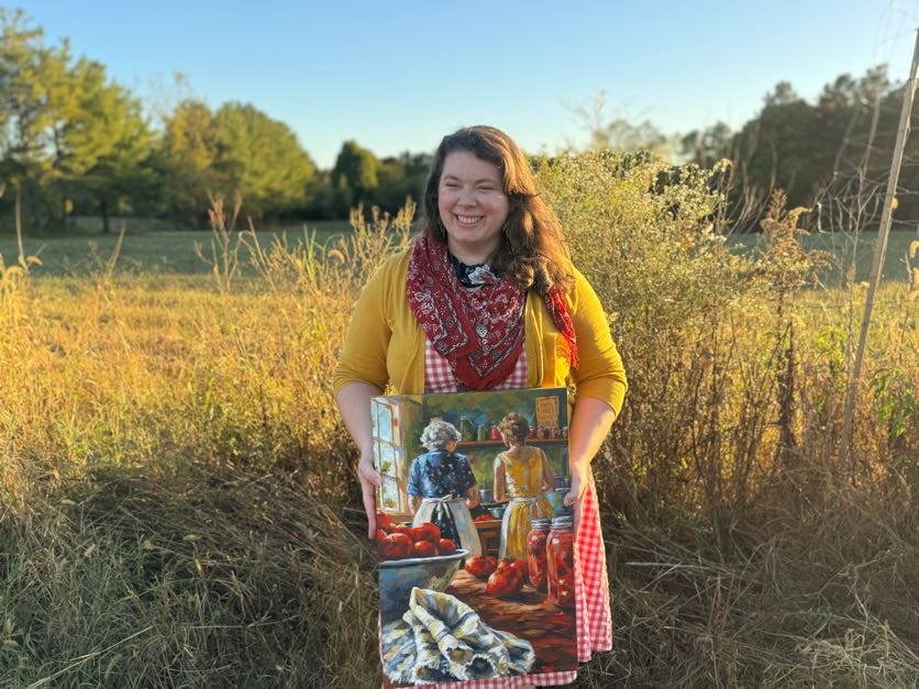 Woman standing outdoors holding a painting of children and pumpkins, with trees and grass in the background.