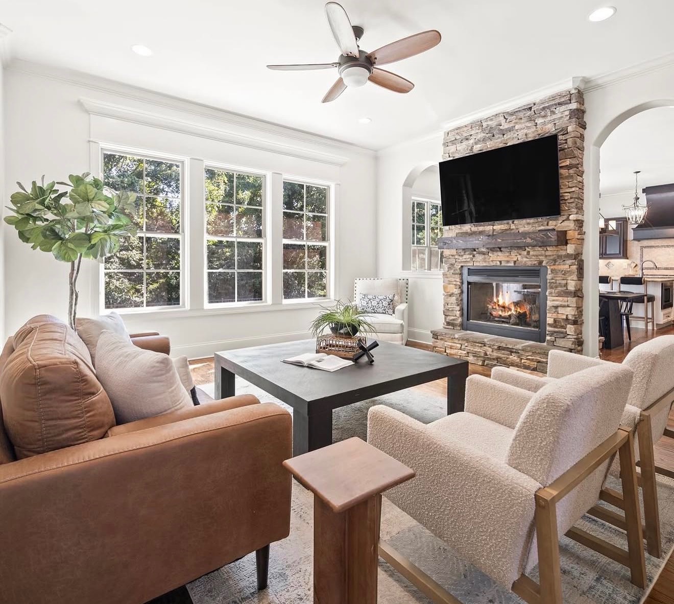 Living room with large windows, a fireplace with a brick surround, a ceiling fan, and seating including a sofa and armchairs.
