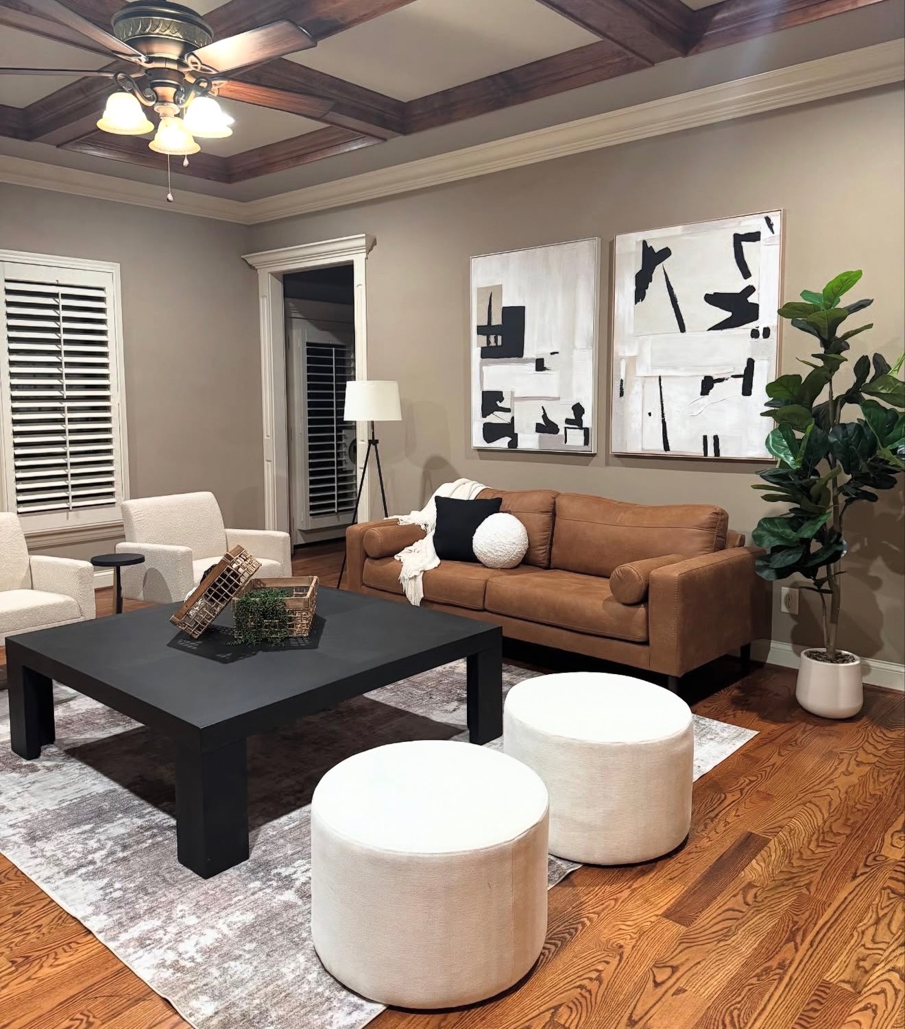 Living room with brown sofa, white ottomans, black coffee table, and wall art, illuminated by ceiling fan and floor lamp.